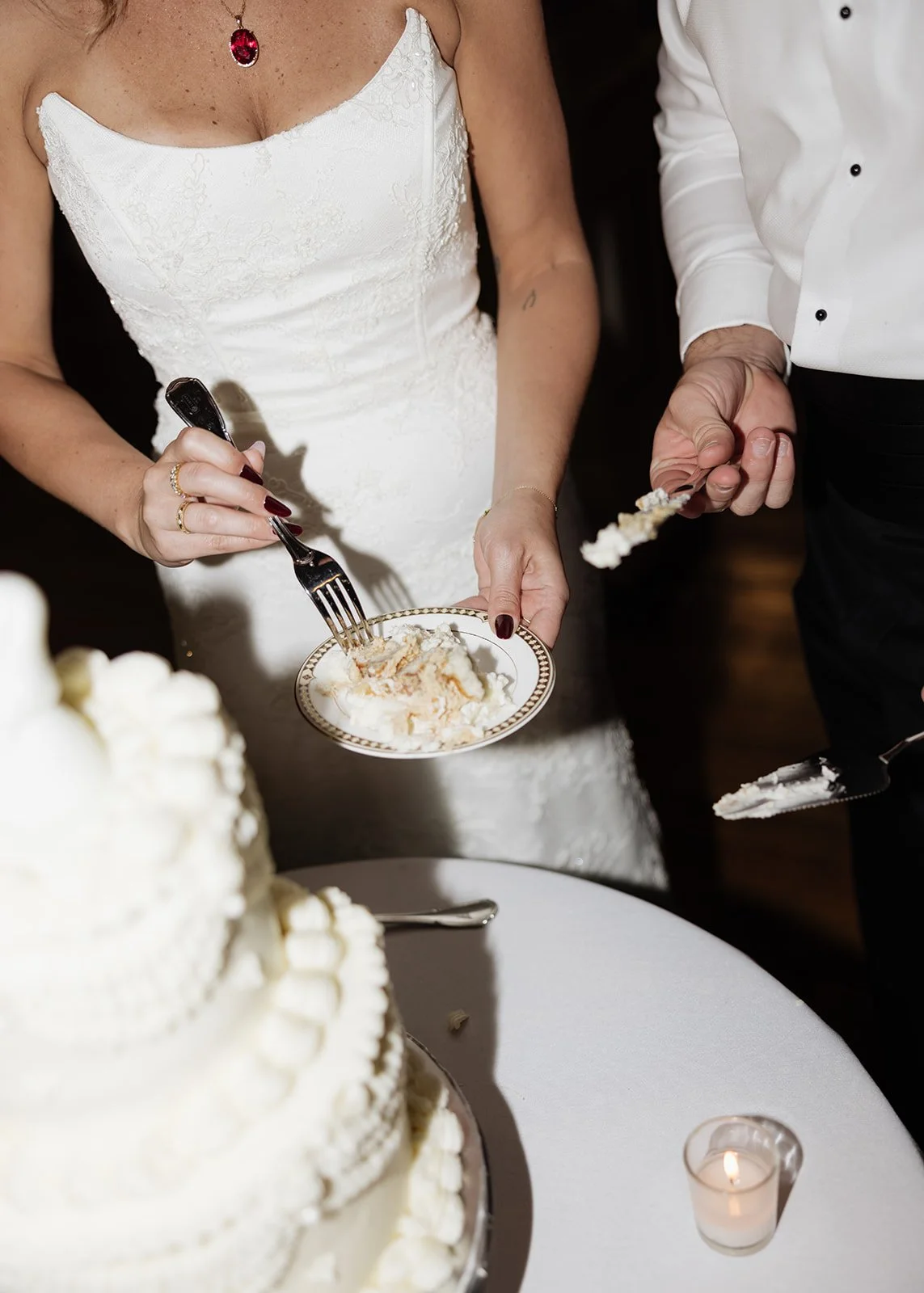 A bride and groom cutting and serving wedding cake. The bride is wearing a white dress and a red gemstone necklace, holding a plate of cake, while the groom, in a white shirt, is holding a piece of cake with a fork.
