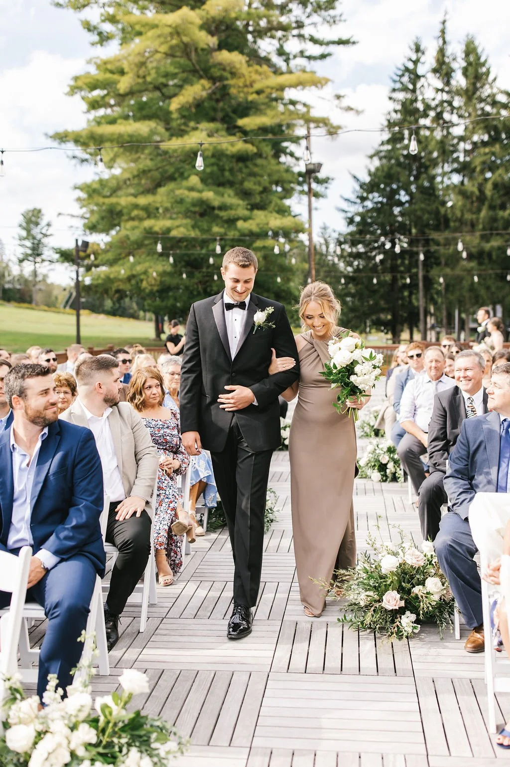 A bride's mother walking her down the aisle at an outdoor wedding ceremony, surrounded by seated guests and decorated with floral arrangements.