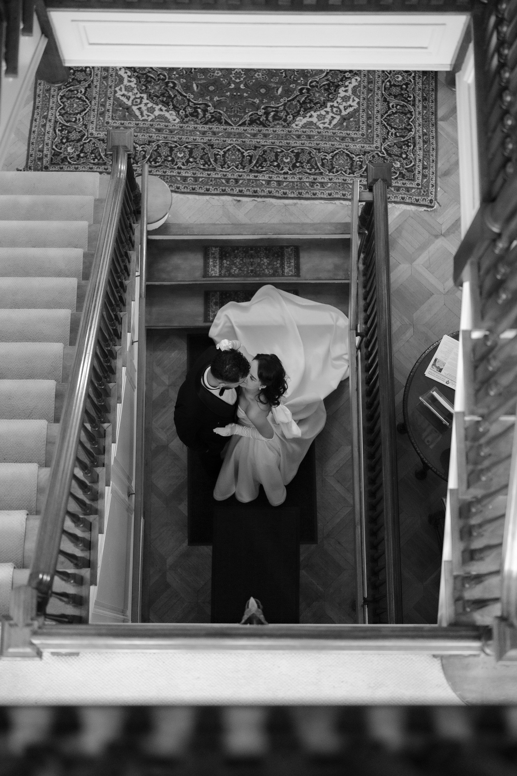 A bride and groom share a kiss on a staircase landing, viewed from above, with a patterned rug and wooden flooring nearby.