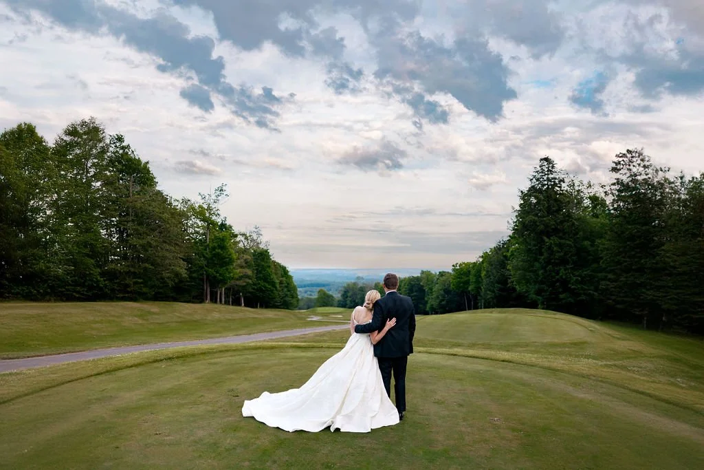 A bride in a white wedding dress and a groom in a dark suit walk arm-in-arm on a grassy path in a spacious park or golf course, surrounded by trees and under a partly cloudy sky.