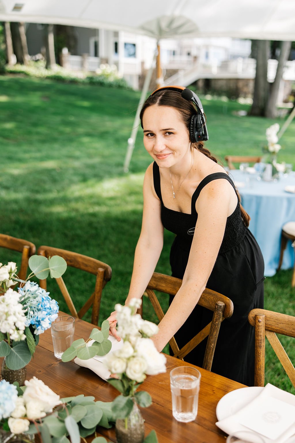 A woman wearing a black dress and a headset is preparing a table outdoors at a garden party or wedding reception, with a wooden table set with flowers, glasses, and plates under a white tent.