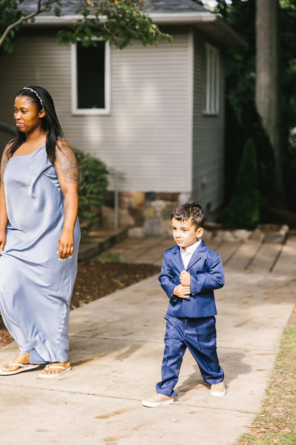 A woman in a light blue dress and a young boy in a blue suit walk outside near a house with gray siding. The woman is smiling while the boy looks ahead with a serious expression.