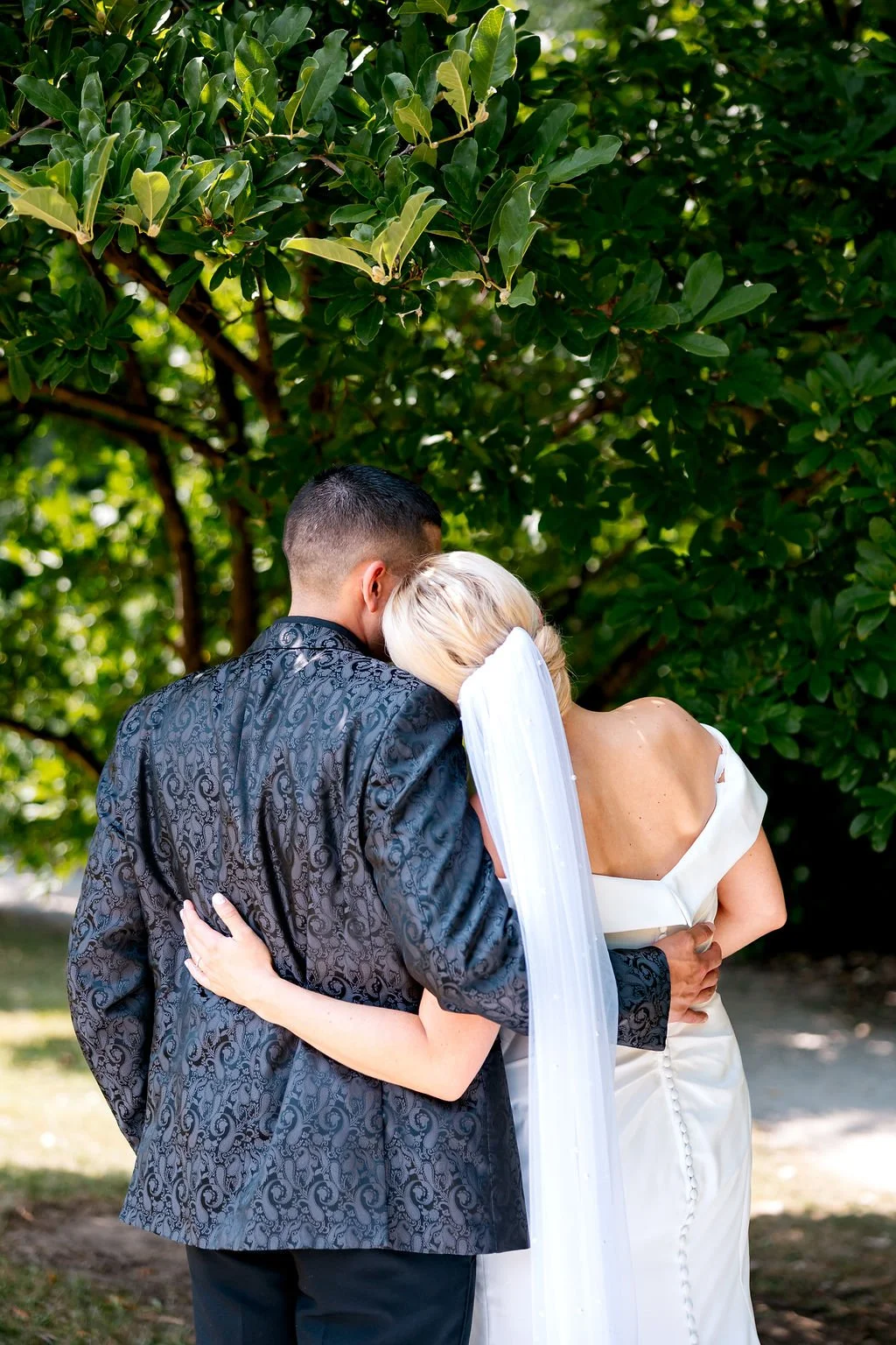A newlywed couple shares a kiss outdoors, holding each other closely with trees and greenery in the background.