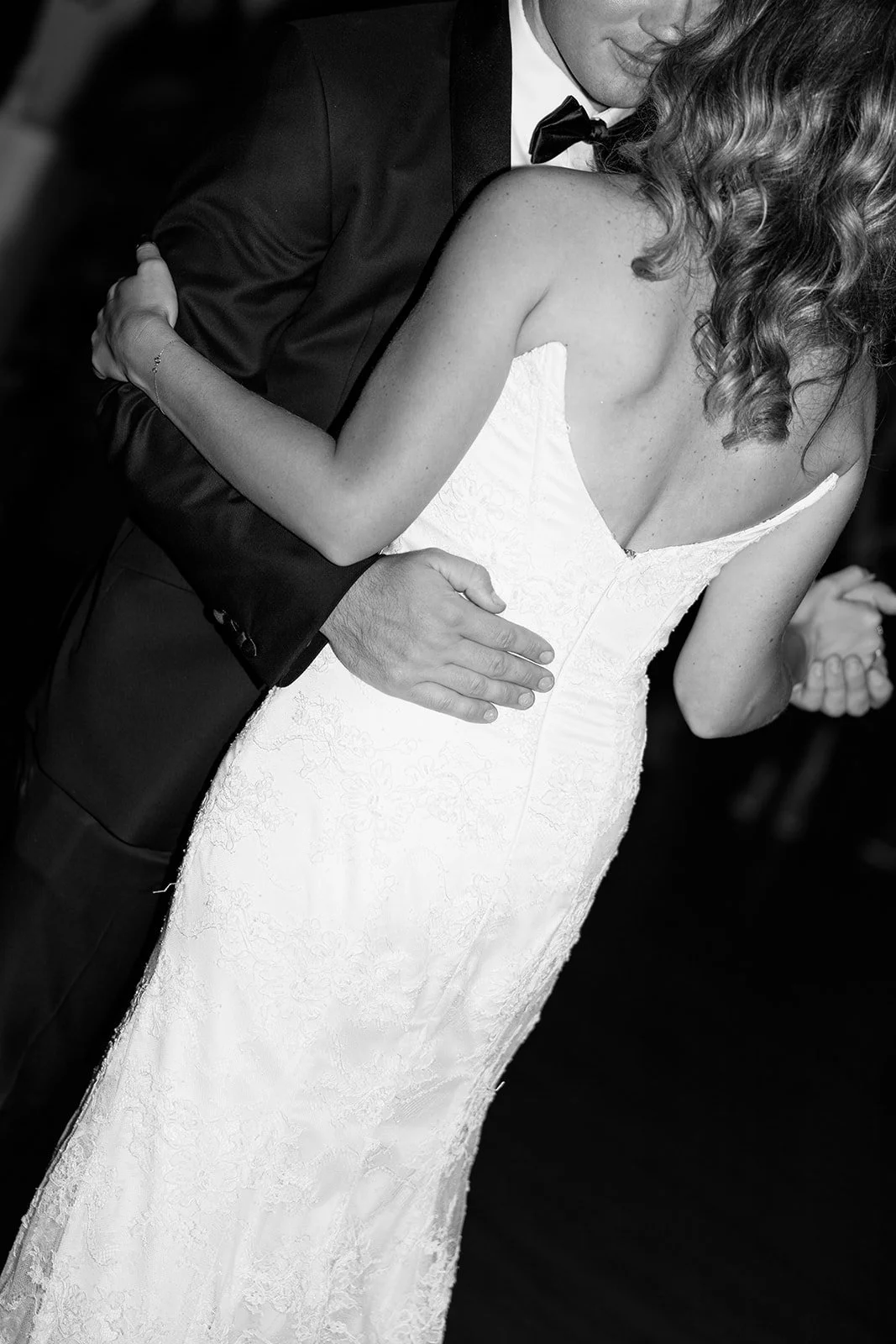 A couple dancing closely at a wedding, with the woman in a white lace dress and the man in a tuxedo, shown in black and white.