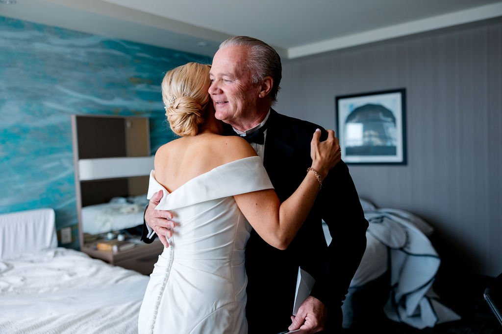 A woman in a white off-shoulder dress hugging an older man in a black tuxedo, both smiling, in a hotel room with unmade beds and a framed picture on the wall.