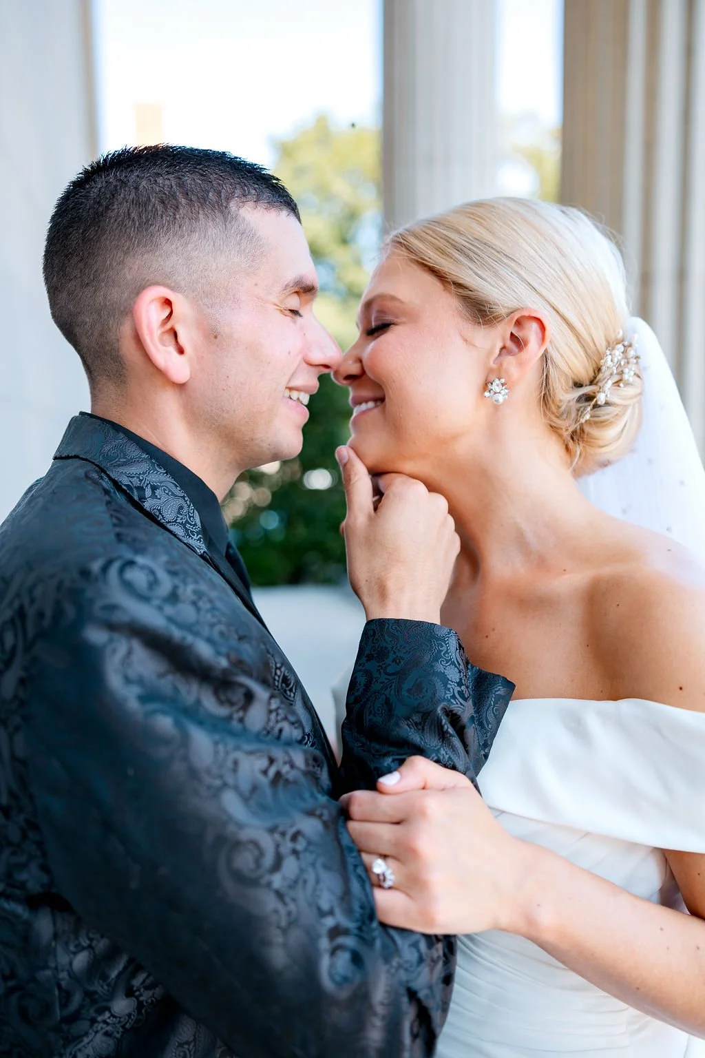 A bride and groom are smiling close to each other, touching noses indoors with a window and greenery in the background.