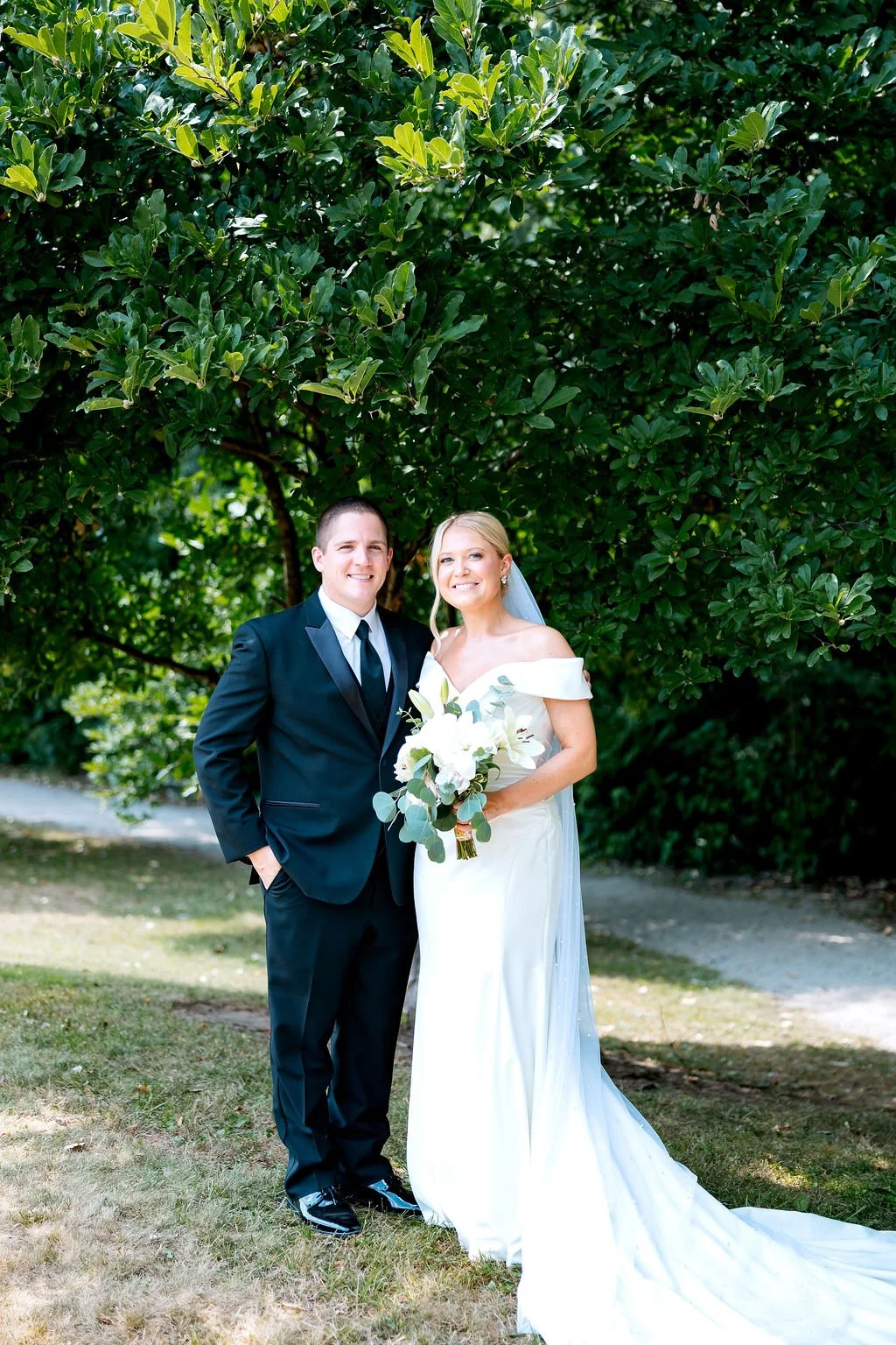 A bride and groom standing outdoors in front of green trees, smiling at the camera. The bride wears a white off-shoulder wedding dress and holds a bouquet of white flowers, while the groom wears a black tuxedo with a white shirt and black tie.