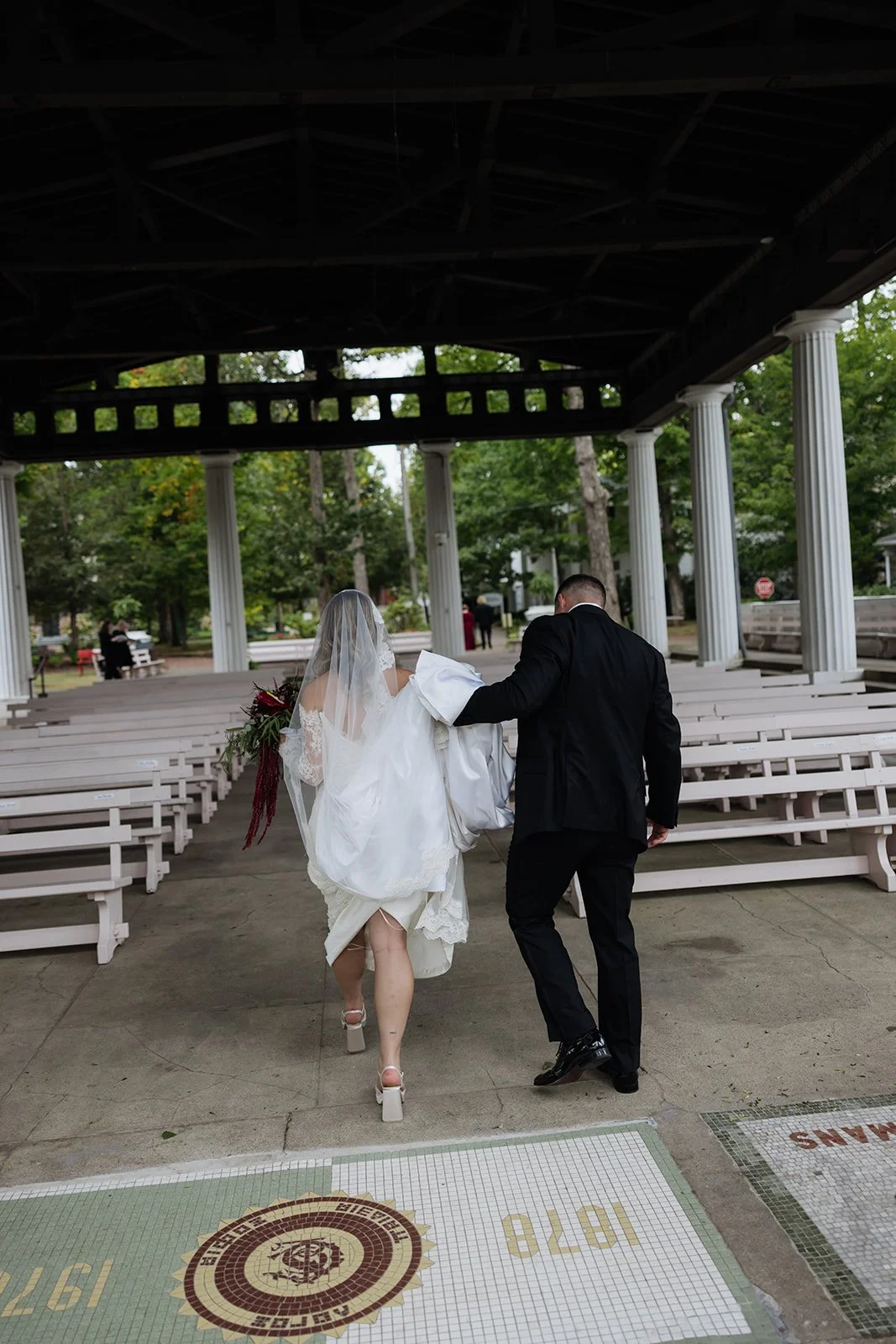 A bride and groom walking away from a wedding ceremony under a gazebo with white benches and pillars, trees in the background, bride holding a bouquet, groom in a black suit, bride in a white wedding dress with veil, the groom helping the bride with 