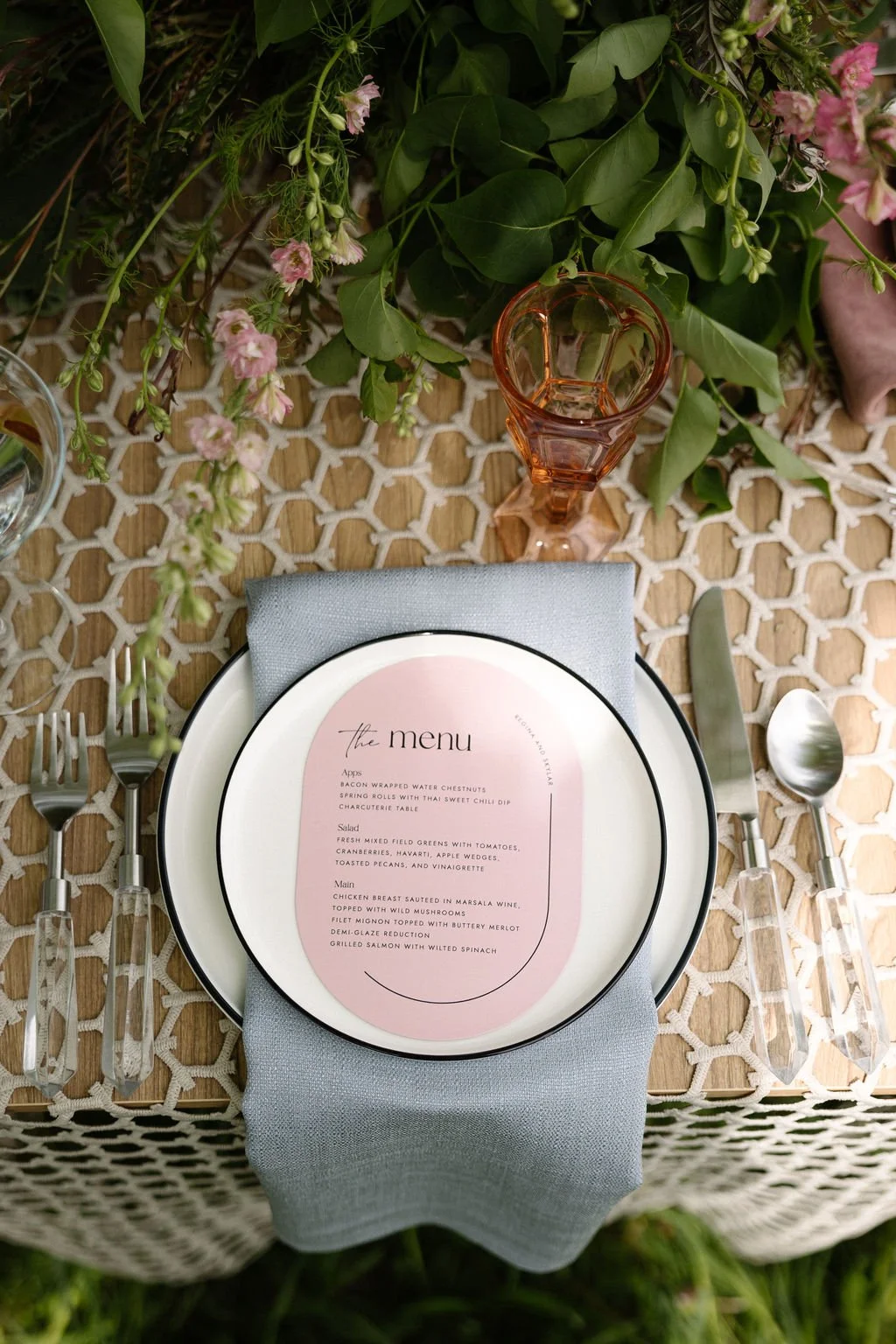 A table setting with a white plate, a blue-gray napkin, and a printed menu. Silverware is placed on either side of the plate. There are green leaves and pink flowers in the background, and a small orange glass. The table has a woven surface.