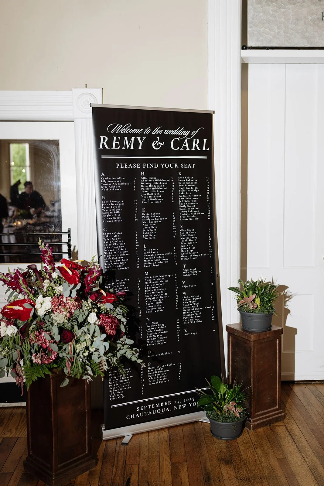 Wedding seating chart at an event, titled 'Welcome to the wedding of Remy & Carl,' with a list of guest names organized alphabetically. The chart is black with white text and is flanked by floral arrangements on wooden stands. The event date is Septe