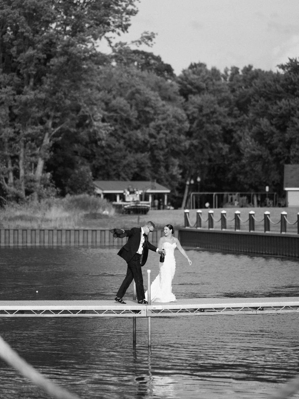 A black and white photo of a bride and groom in wedding attire dancing on a floating dock on a lake, with trees and houses in the background.