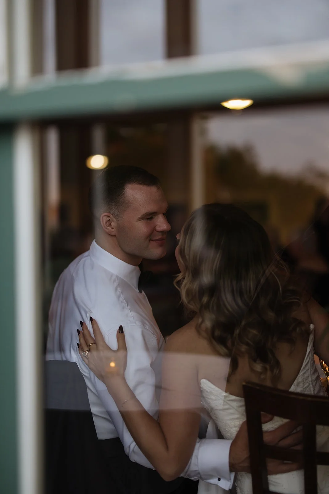 A couple sharing a romantic moment seen through a window, with the man smiling gently and the woman with long wavy hair.