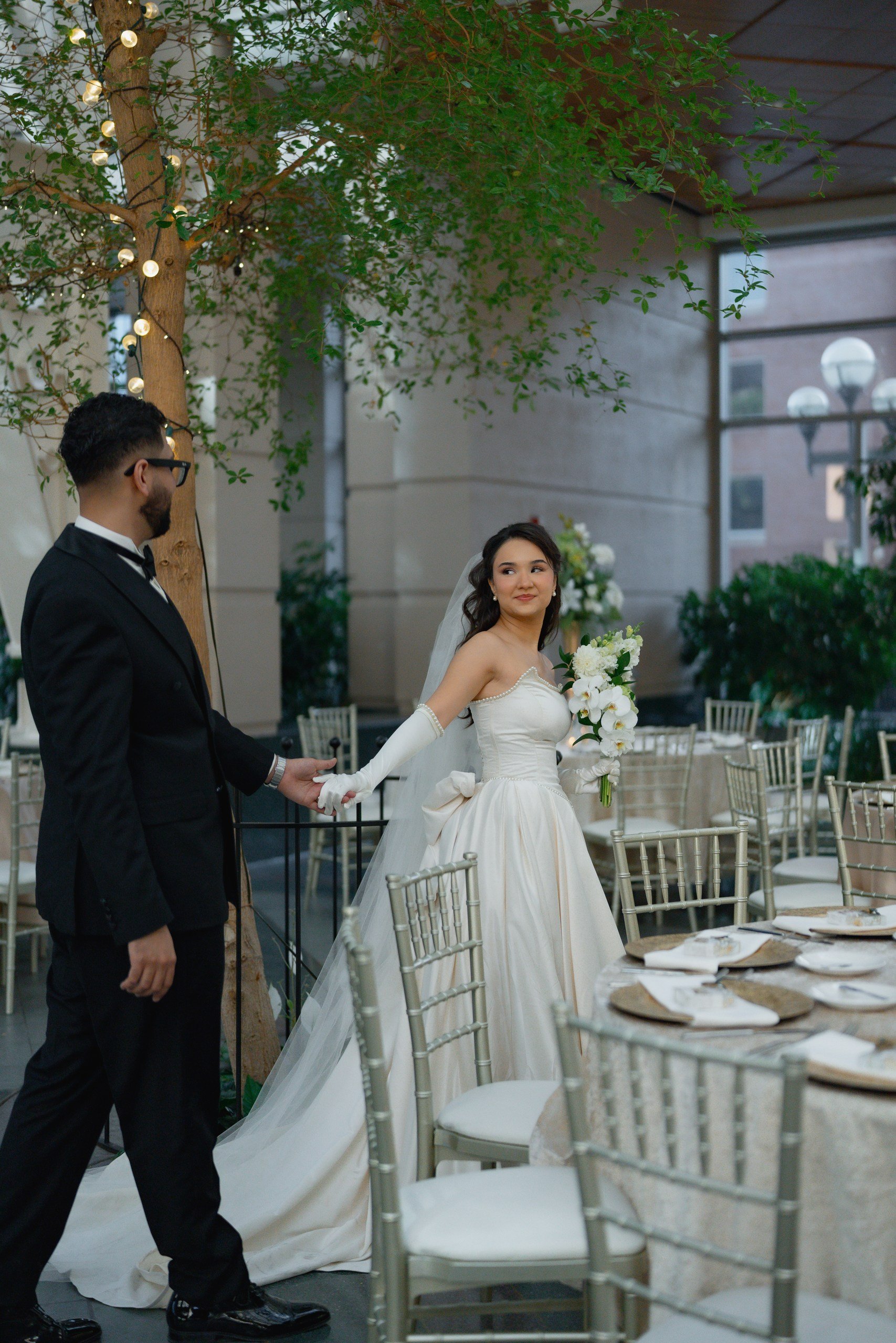 A bride and groom holding hands, inside a decorated venue with tables and chairs, a large tree with leaves and string lights, and a window showing a brick building outside.