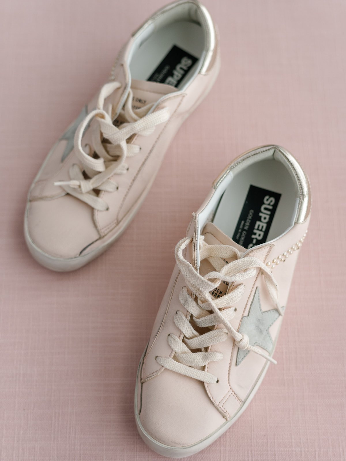 Pair of white sneakers with metallic gold accents and star designs on a pink textured surface.
