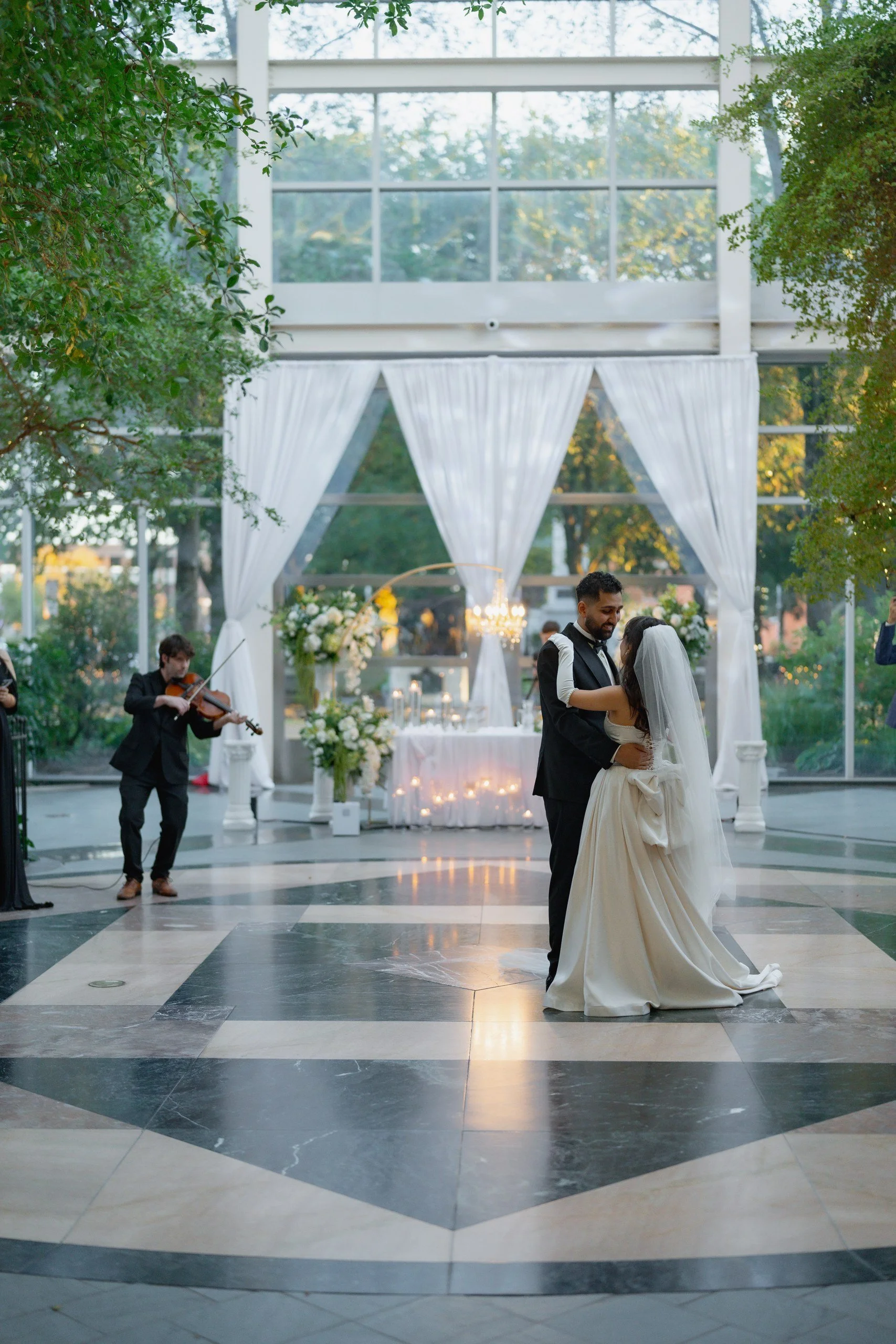 A bride and groom share a dance during their wedding reception in an elegant, open-air indoor venue decorated with white drapes and flowers. A violinist performs nearby, and lush greenery can be seen through large windows.