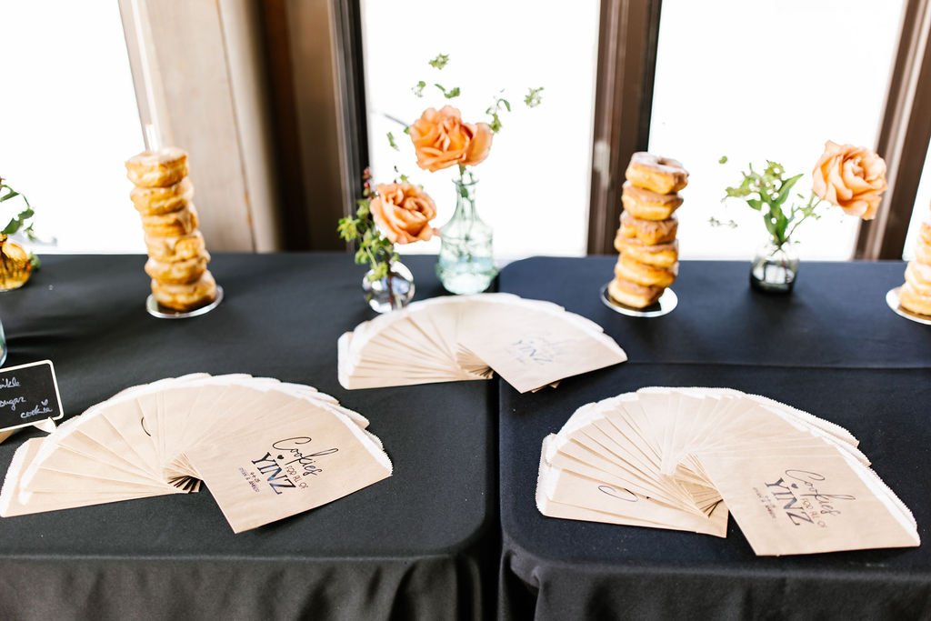 Table with black tablecloth decorated with floral arrangements in glass vases, stacks of fan-shaped cards, and a sign that reads "Table YINZ" for a wedding or special event.
