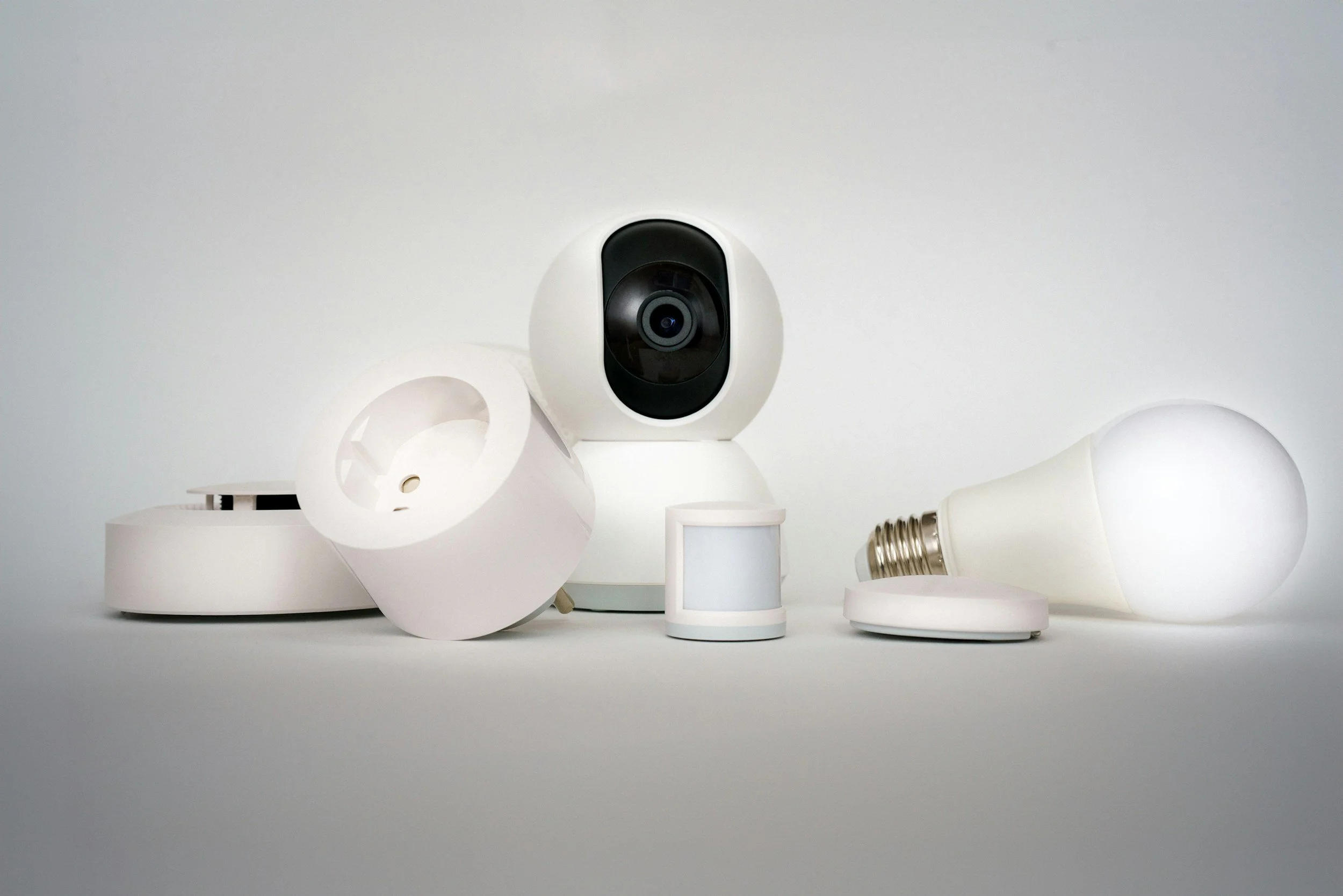 Various white objects including a black security camera, a white lampshade, a light bulb, and other light fixtures arranged on a white surface against a white background.