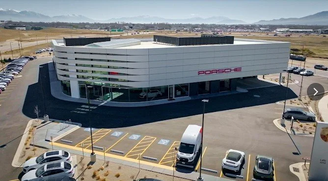 An aerial view of a modern Porsche building with a parking lot in front, located in a semi-rural area with mountains in the background.