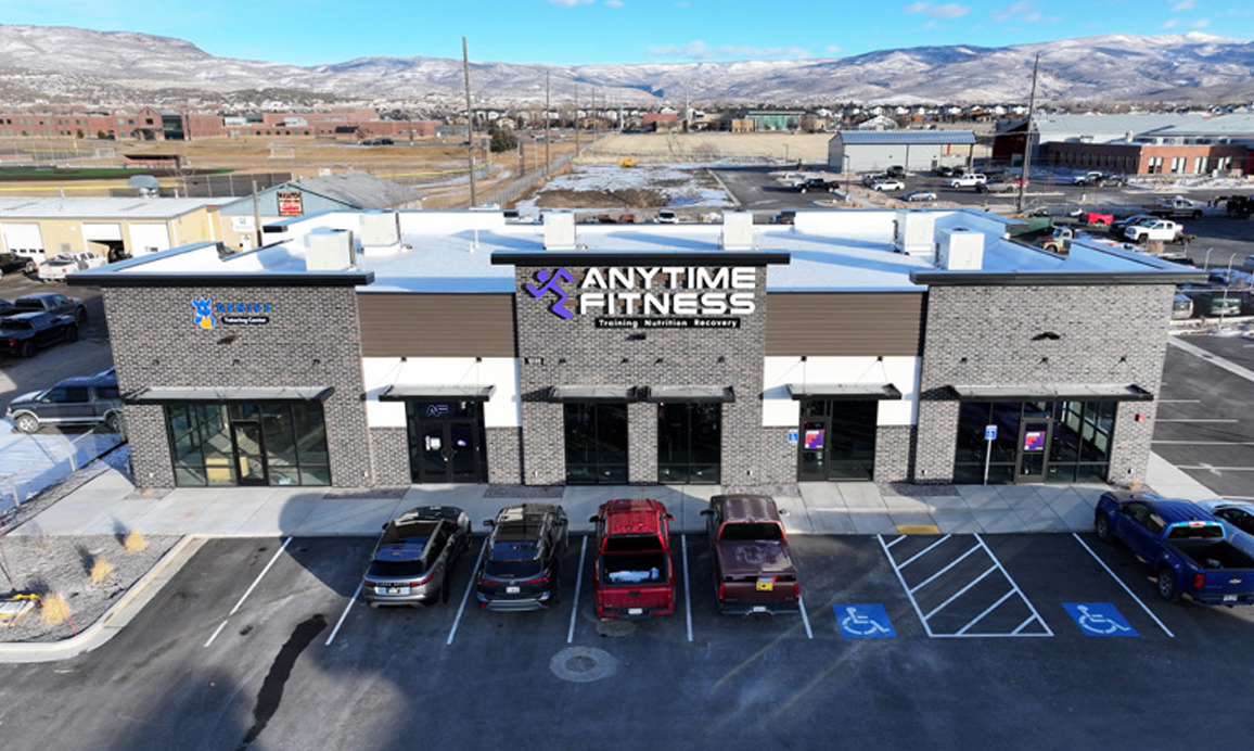 A two-story commercial building with a fitness center named 'Anytime Fitness' with parking spaces, including designated handicapped spots in the front, and a mountainous landscape in the background.