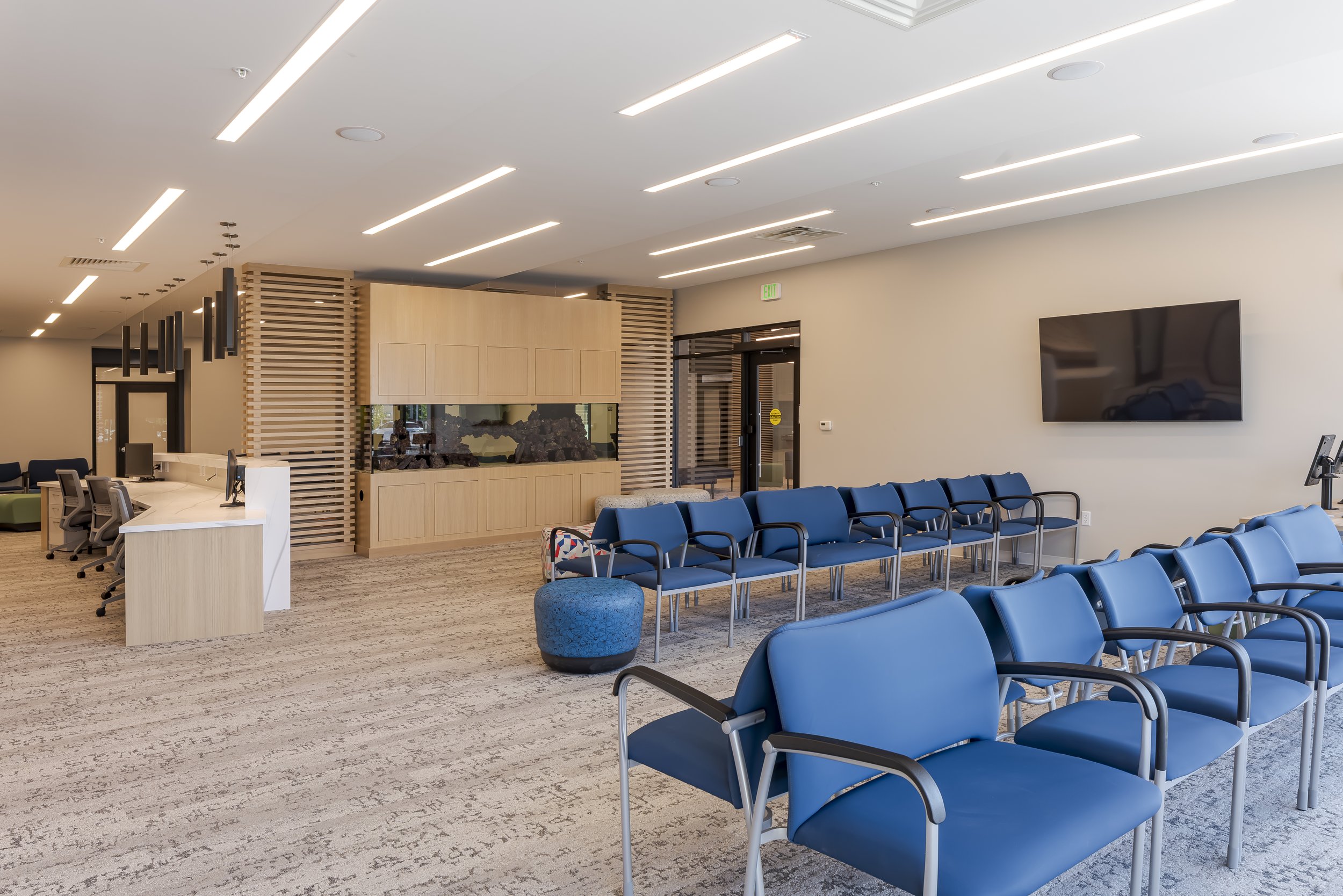 A spacious waiting area with multiple rows of blue chairs, a large flat screen TV on the wall, and a curved reception desk with multiple computer workstations. The room features modern lighting fixtures, a wood-paneled wall with a built-in aquarium, and beige walls with a gray carpet.