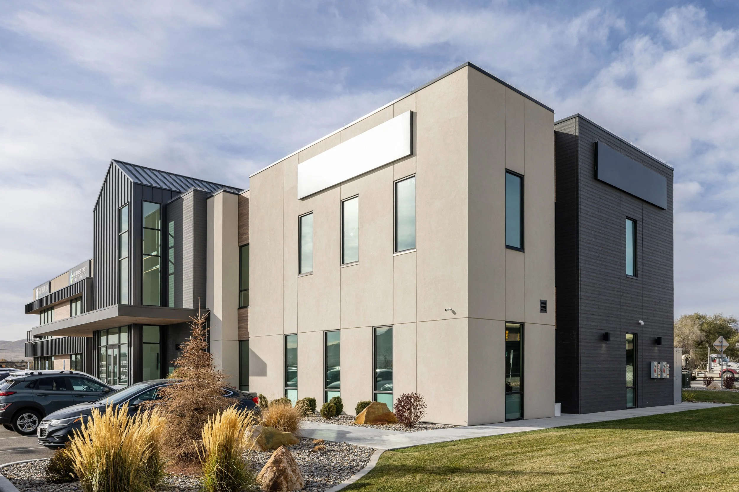 Modern commercial building with large windows, black and beige exterior walls, parking lot, and landscaped front yard with ornamental grasses and rocks.