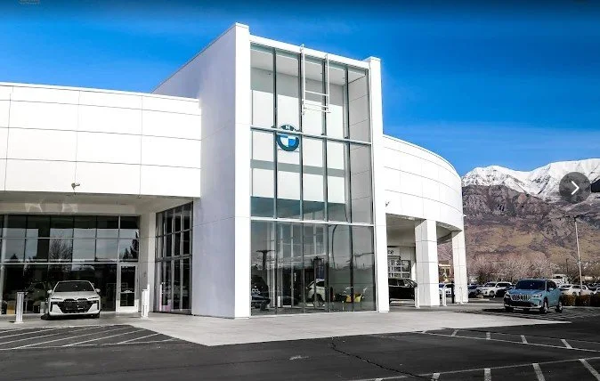 Exterior view of a BMW car dealership with glass windows and white building, parked cars outside, mountains in the background under a blue sky.