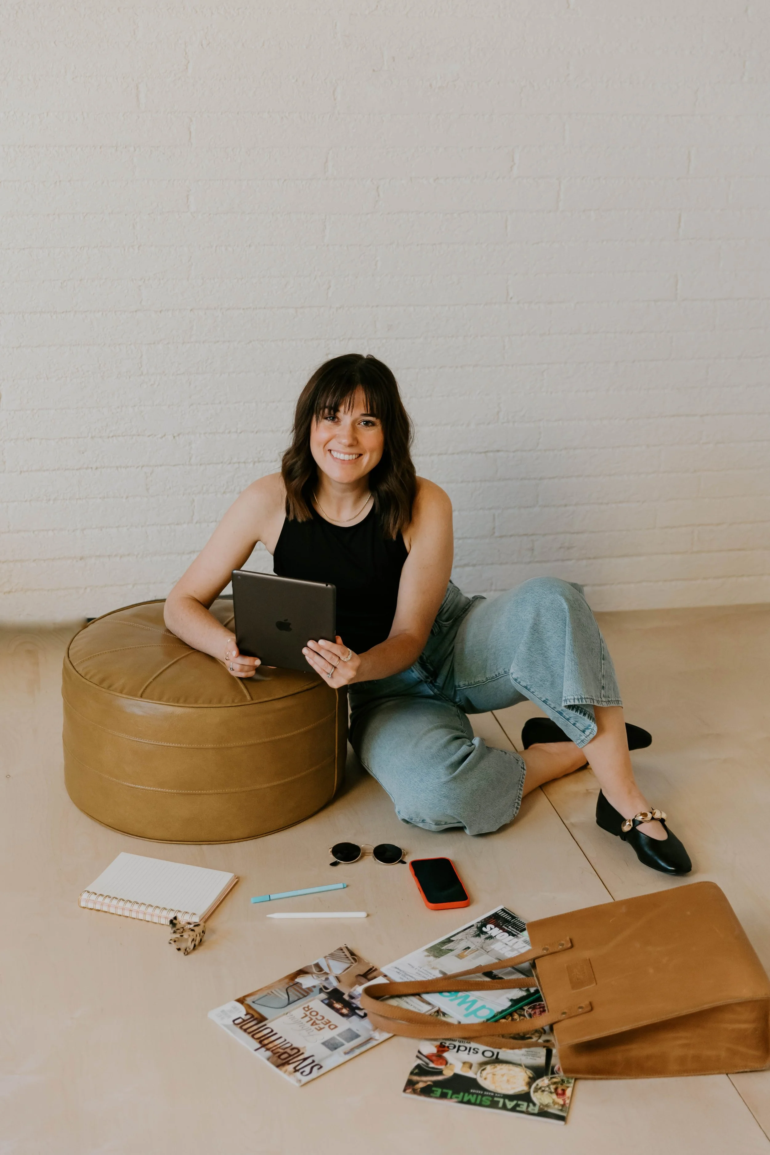 A young woman with dark hair and a black sleeveless top, sitting on the floor, holding a tablet, smiling, with a white textured wall behind her. A tan pouf is next to her, and various items including magazines, a notebook, sunglasses, a red phone, and a brown purse are on the floor.