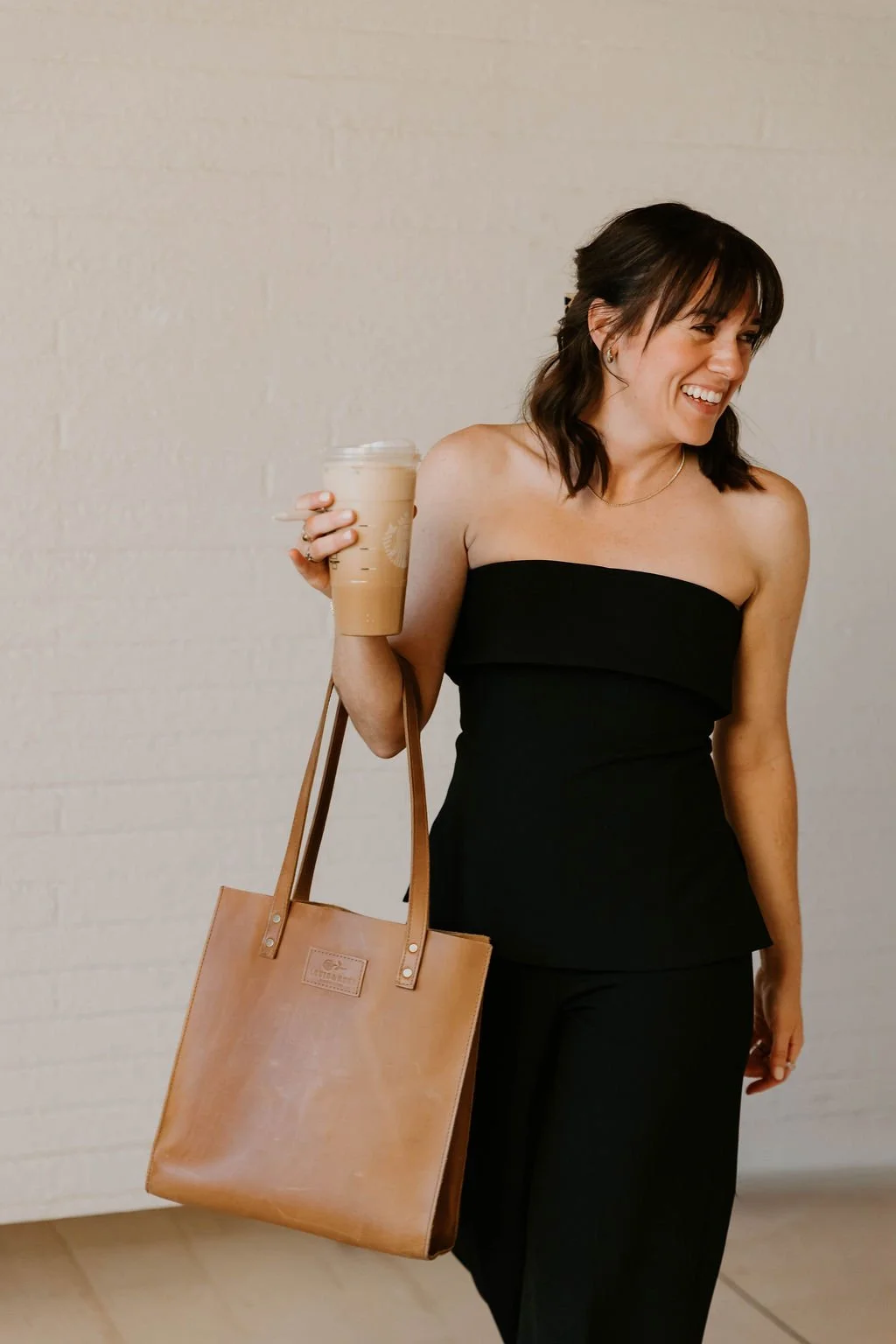Woman smiling, wearing a strapless black dress, holding a large iced coffee, and carrying a tan tote bag, standing against a plain cream-colored wall.