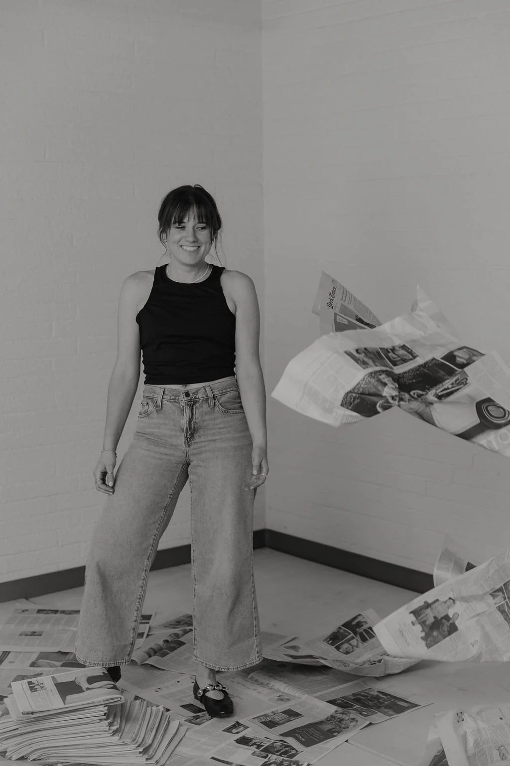 A woman smiling and standing in a room with scattered newspapers on the floor and in mid-air.