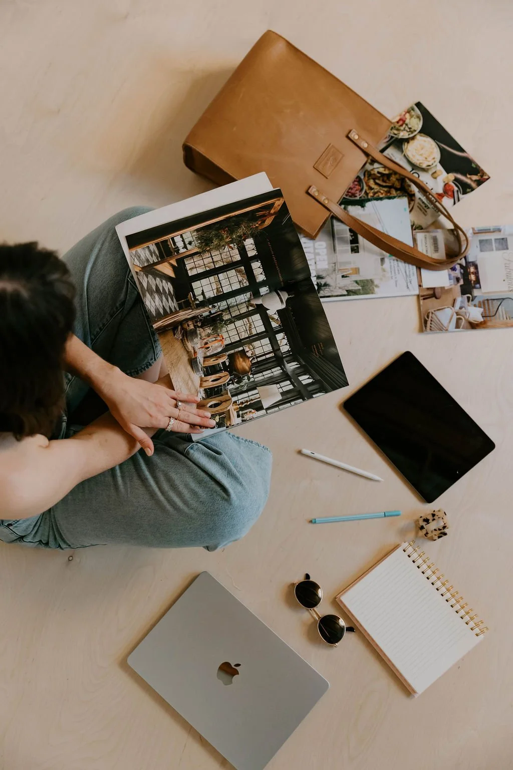 A person sitting on the floor looking at a magazine showing an interior space, surrounded by a laptop, tablet, pens, notes, sunglasses, and a brown leather bag, with magazines and a snack nearby.