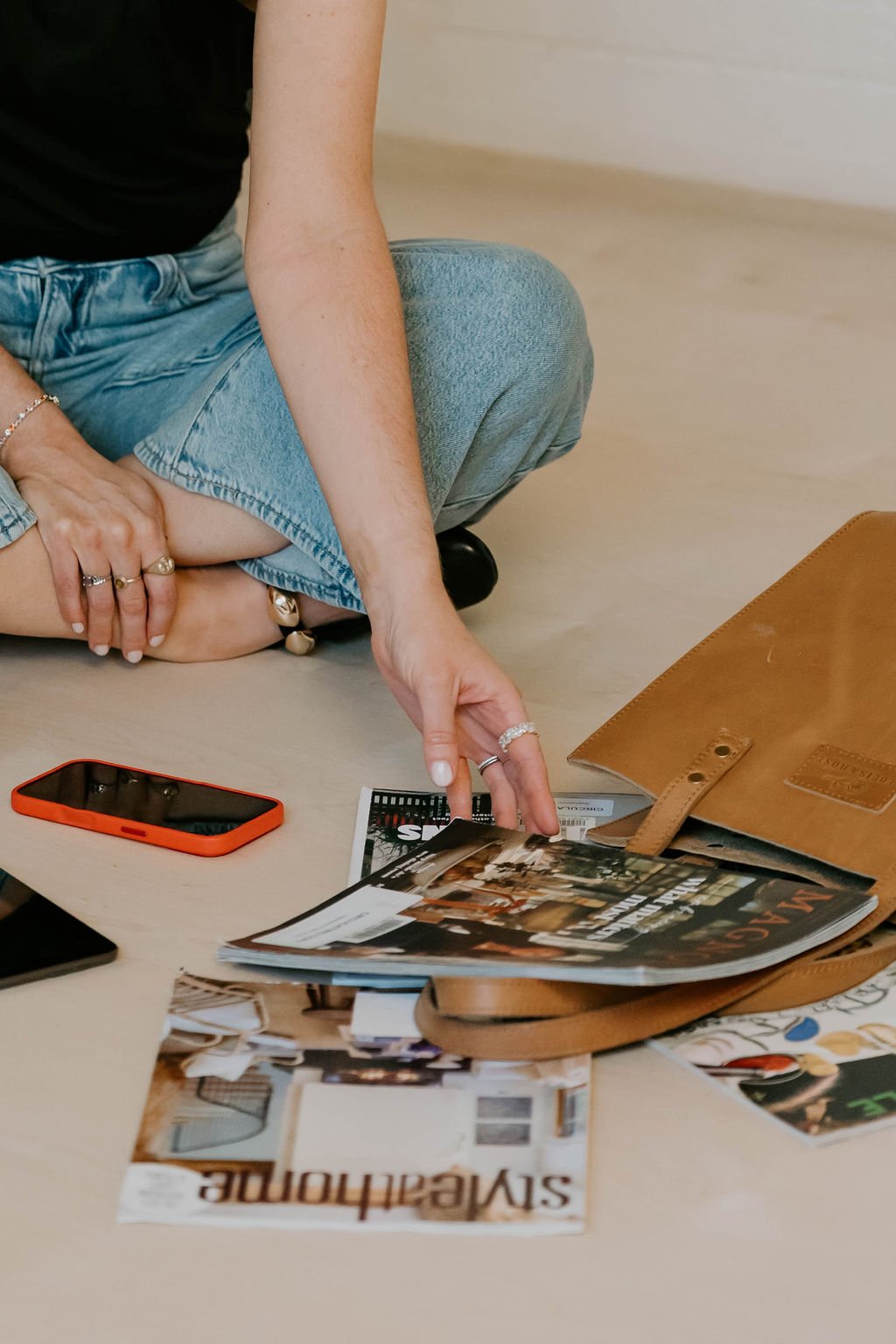 A person is sitting on the floor with a magazine in hand, surrounded by magazines, a brown leather bag, a smartphone, and other items.