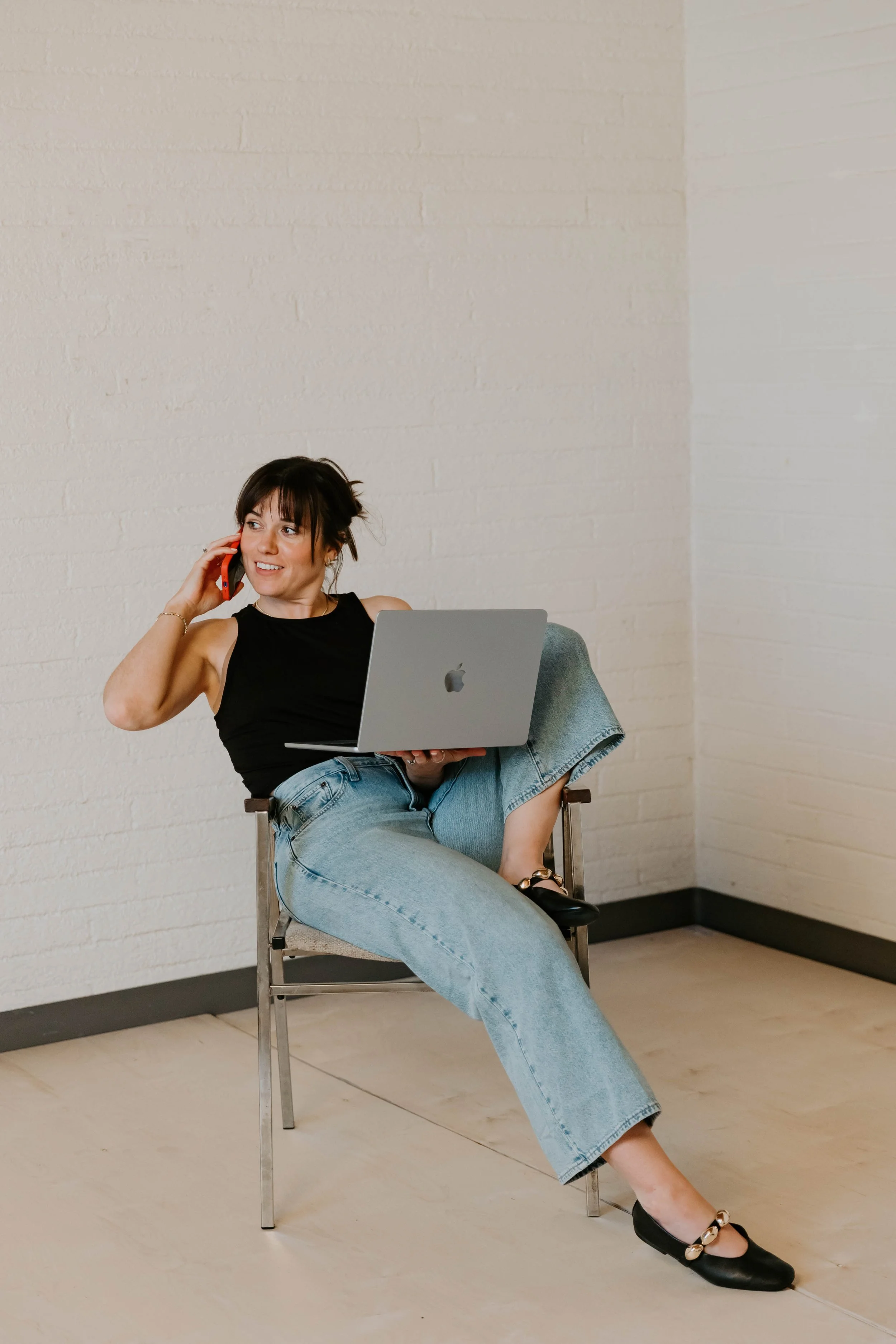 A woman sitting on a chair with her legs crossed, holding a laptop on her lap, talking on a phone with a smile, wearing a black sleeveless top and blue jeans, in a room with white brick walls.