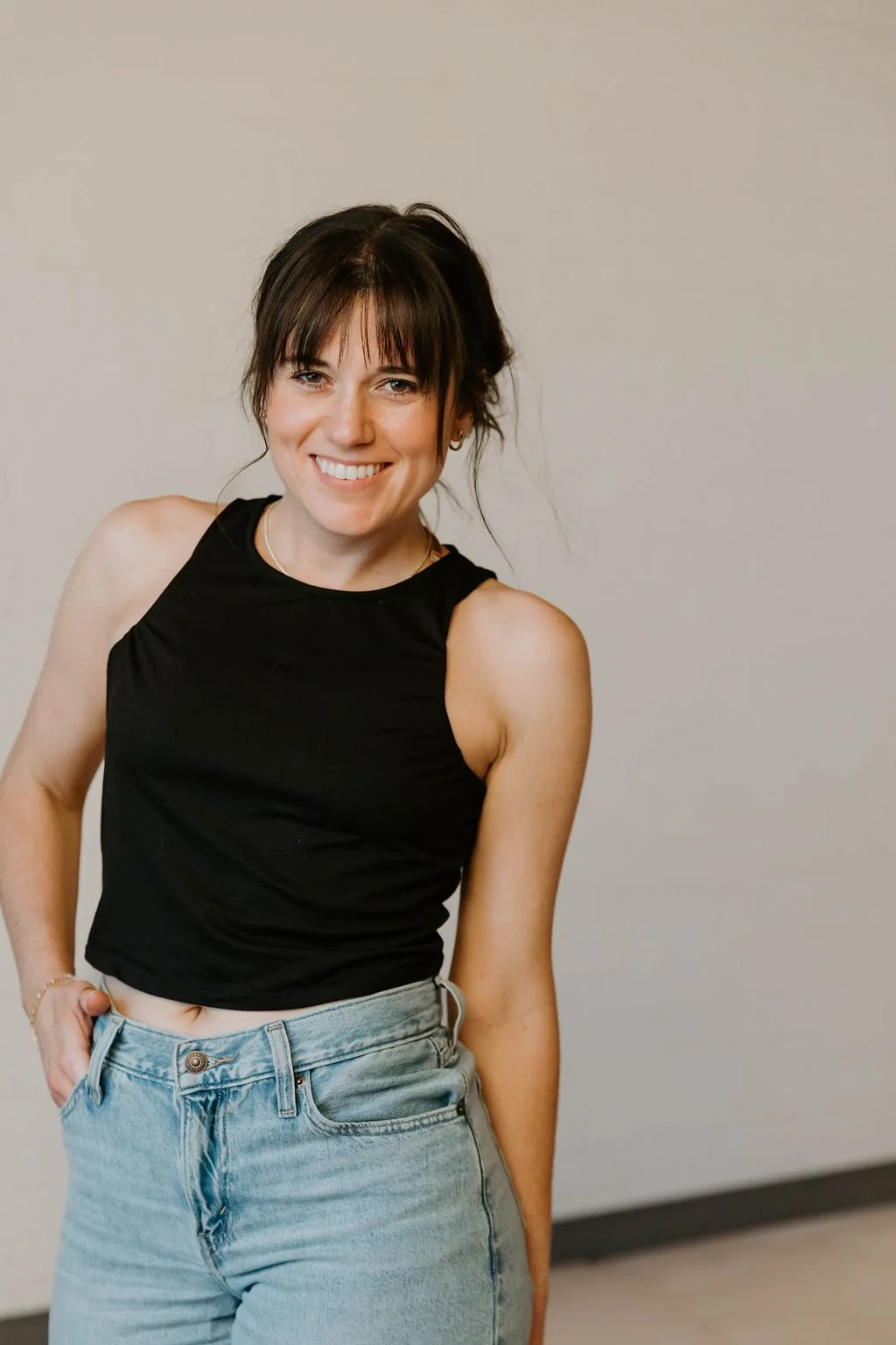 A woman with short dark hair smiling at the camera, wearing a black sleeveless top and light blue jeans, standing against a plain wall.