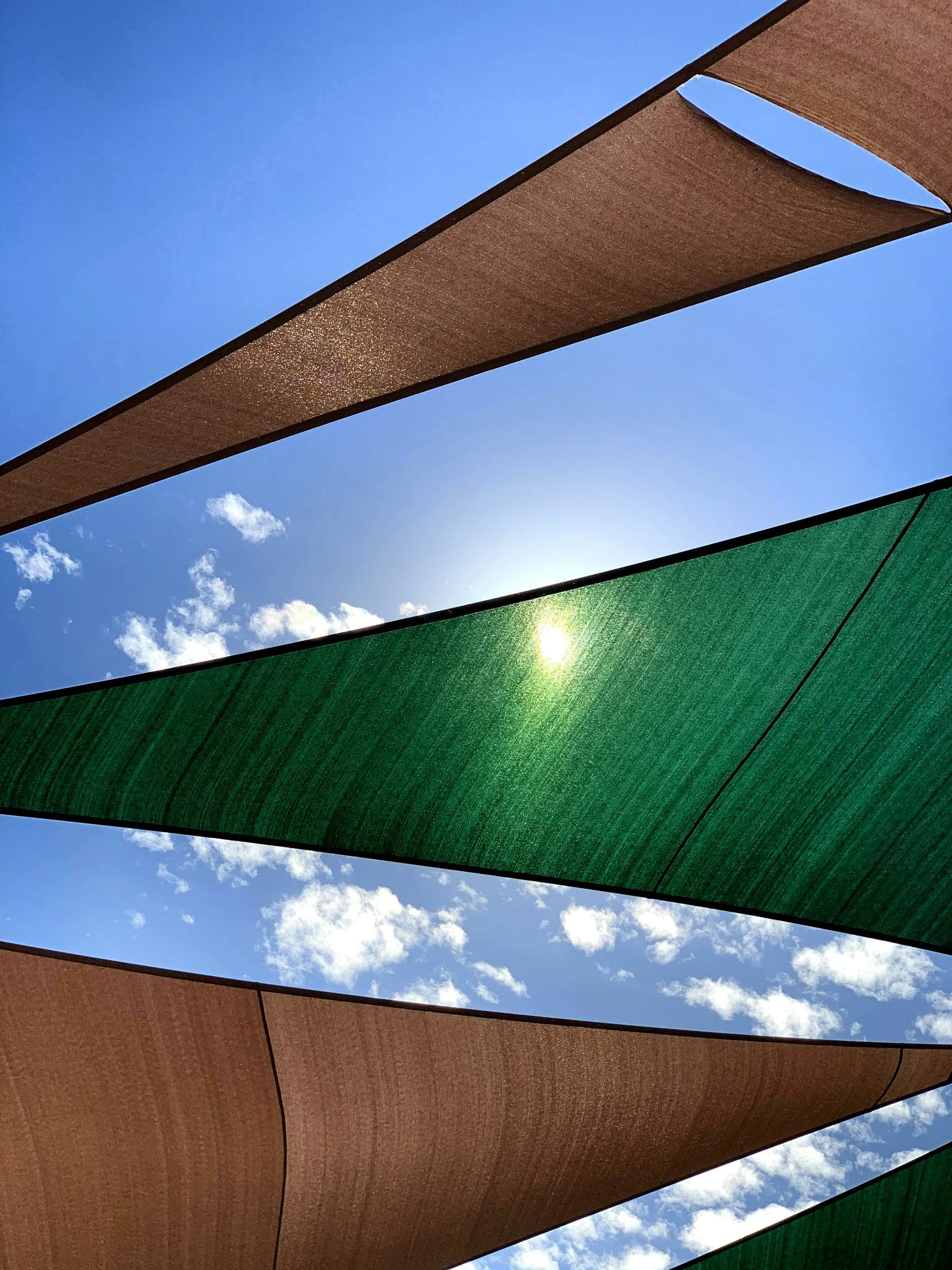 Colorful fabric shades in blue, brown, and green, stretched outdoors under a partly cloudy sky.