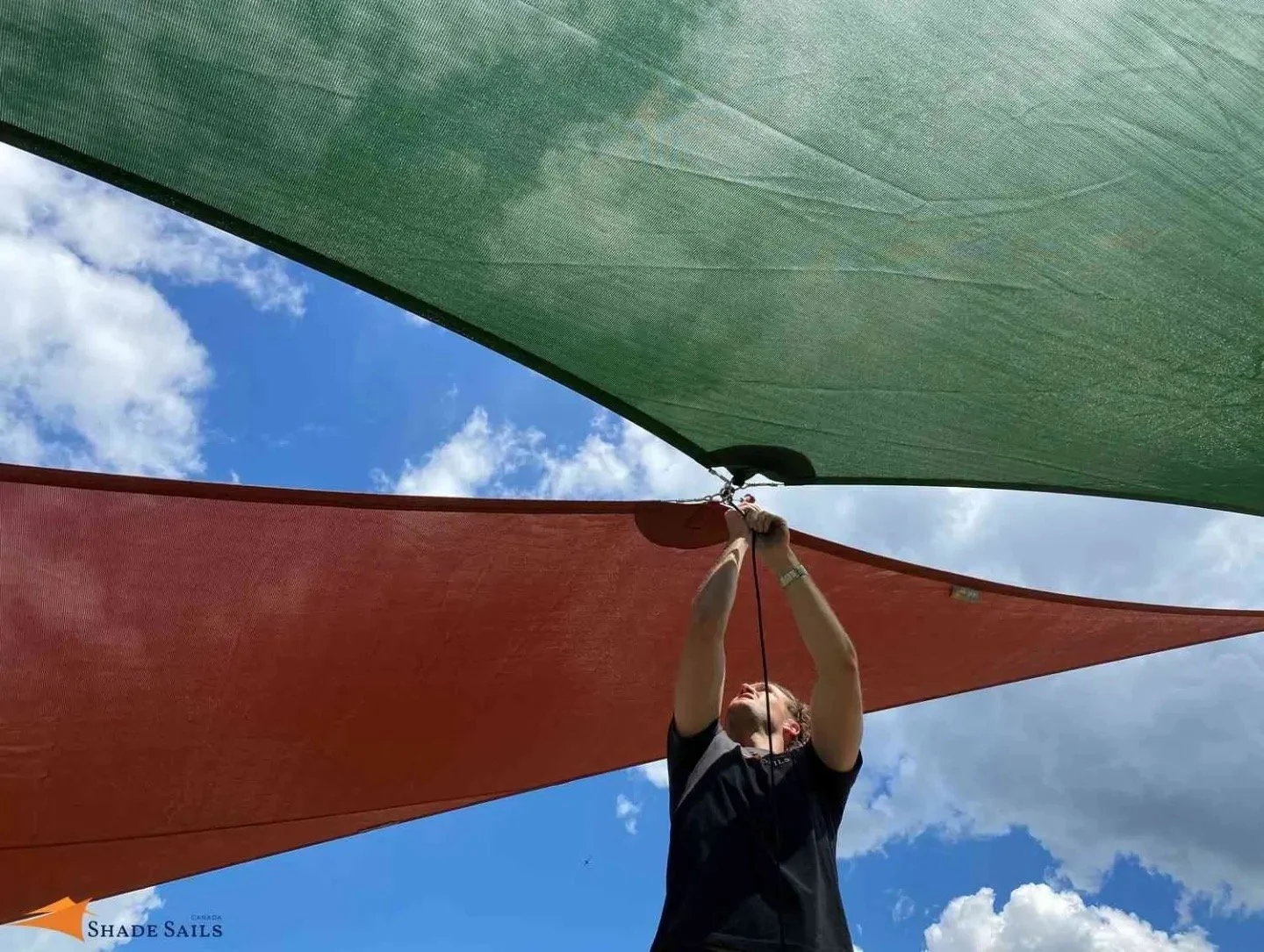 Person adjusting or installing green and red sail shades outdoors, with a blue sky and white clouds in the background.