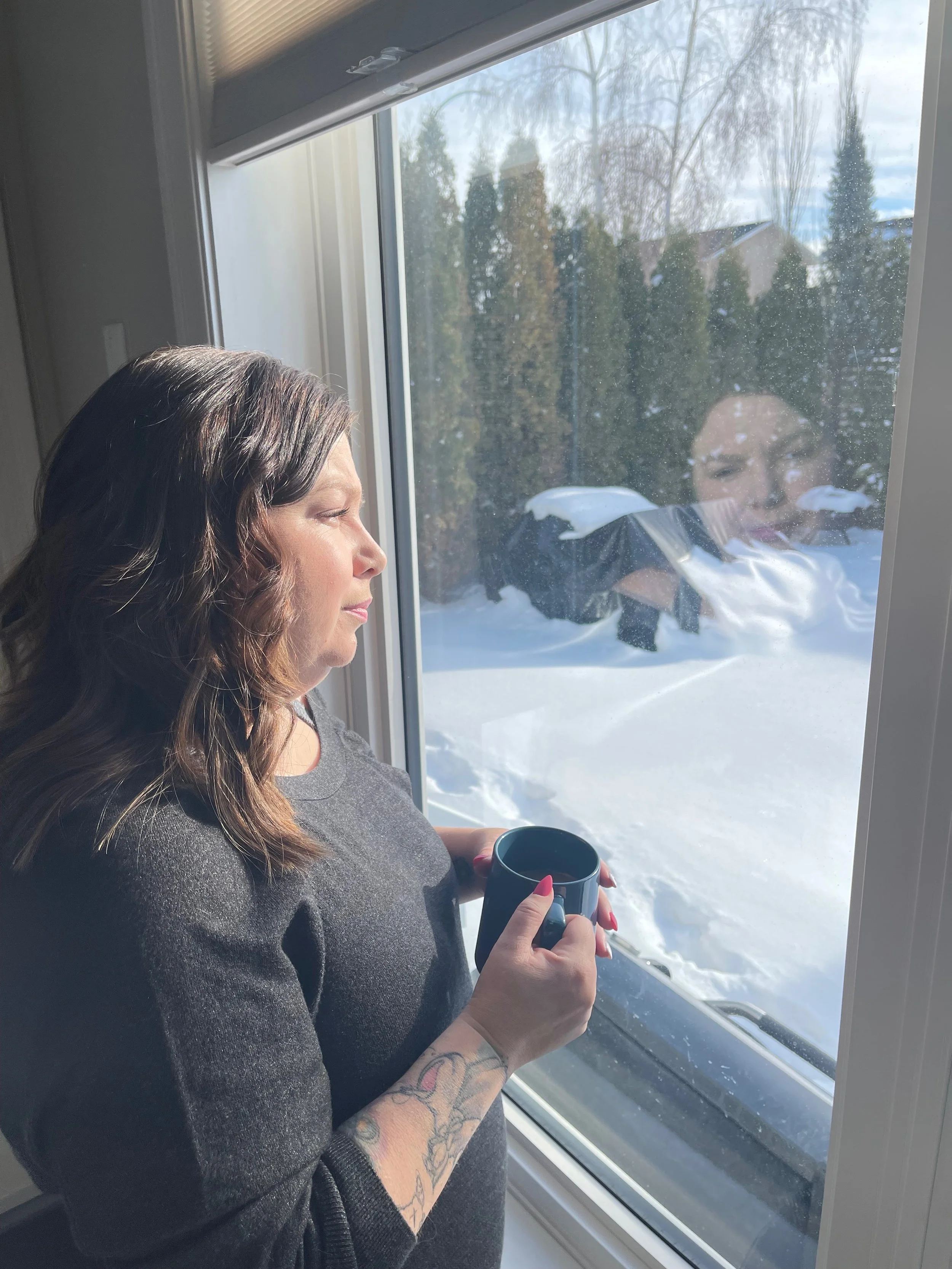 A woman with long, wavy brown hair and tattooed arm, holding a coffee mug, looks out of a window at a snowy outdoor scene with trees and houses.