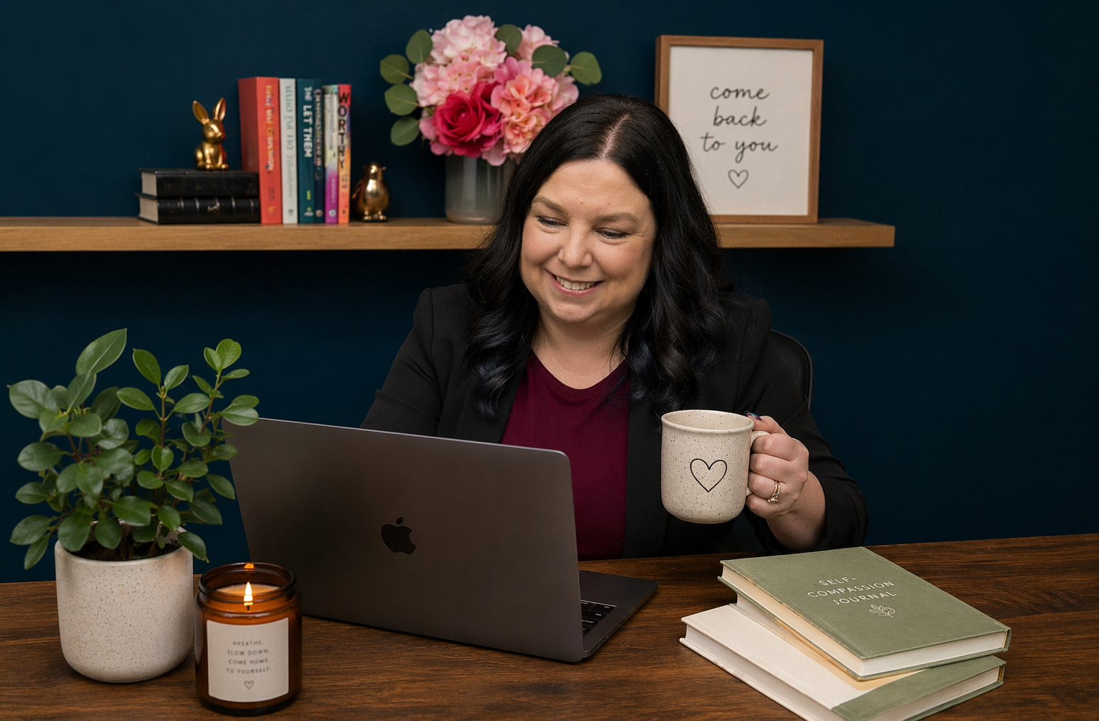 A woman with dark hair sits at a wooden desk working on a laptop, holding a mug with a heart design. The desk has books, a plant, and a candle. Behind her, a shelf with colorful books, a flower arrangement, and a framed sign that says 'come back to you' is visible.