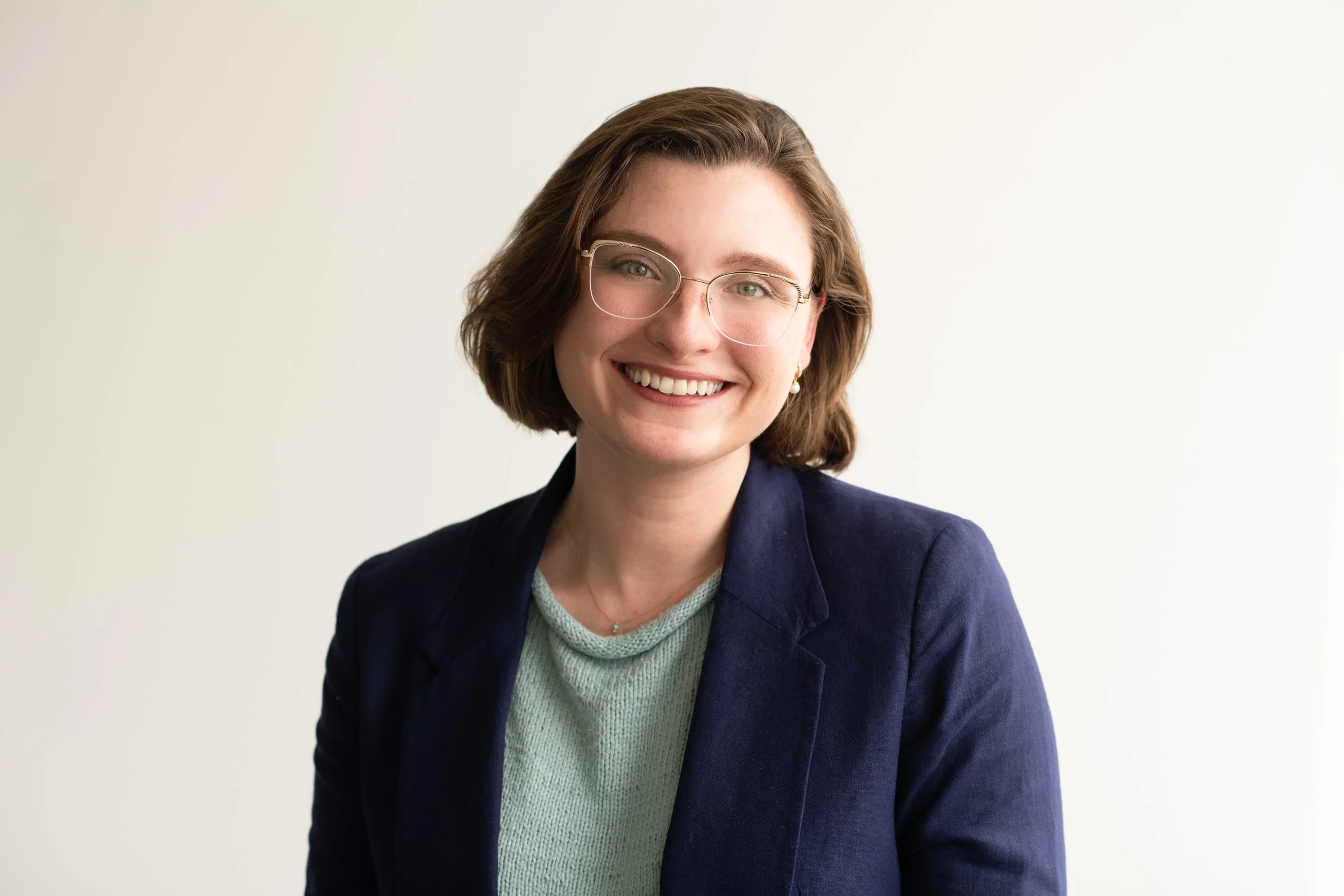 Portrait of a smiling woman with short brown hair, glasses, wearing a navy blazer and a light-colored sweater, against a plain white background.