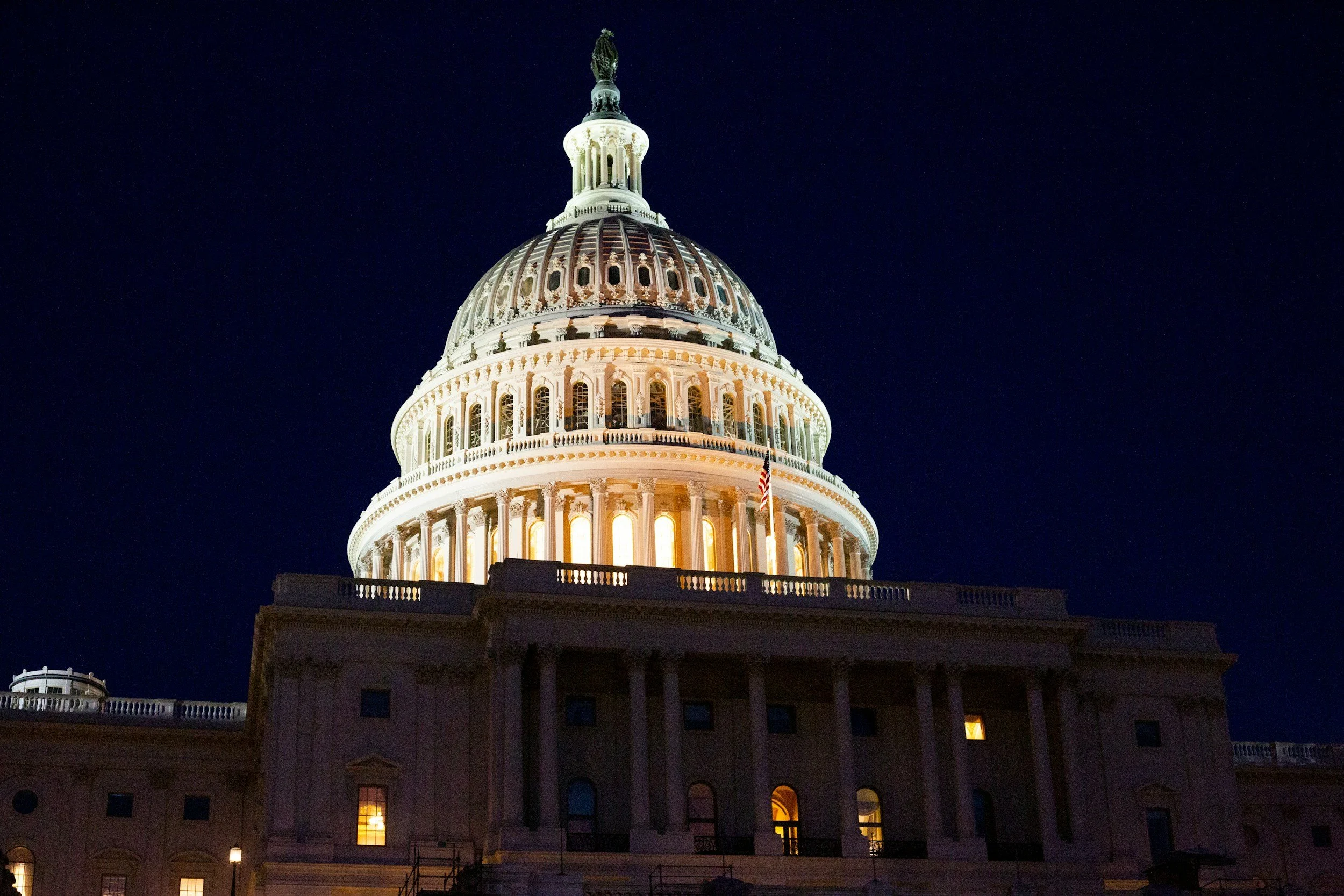 Nighttime view of the United States Capitol building with its dome illuminated, showing the statue on top, against a dark sky with stars.