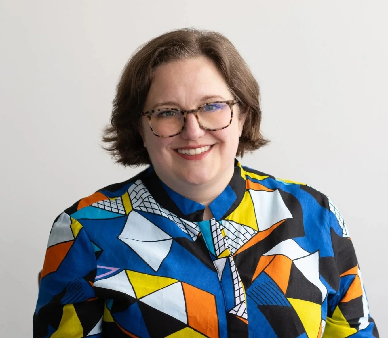 A woman with short brown hair and glasses smiling, wearing a colorful geometric-patterned jacket against a plain white background.