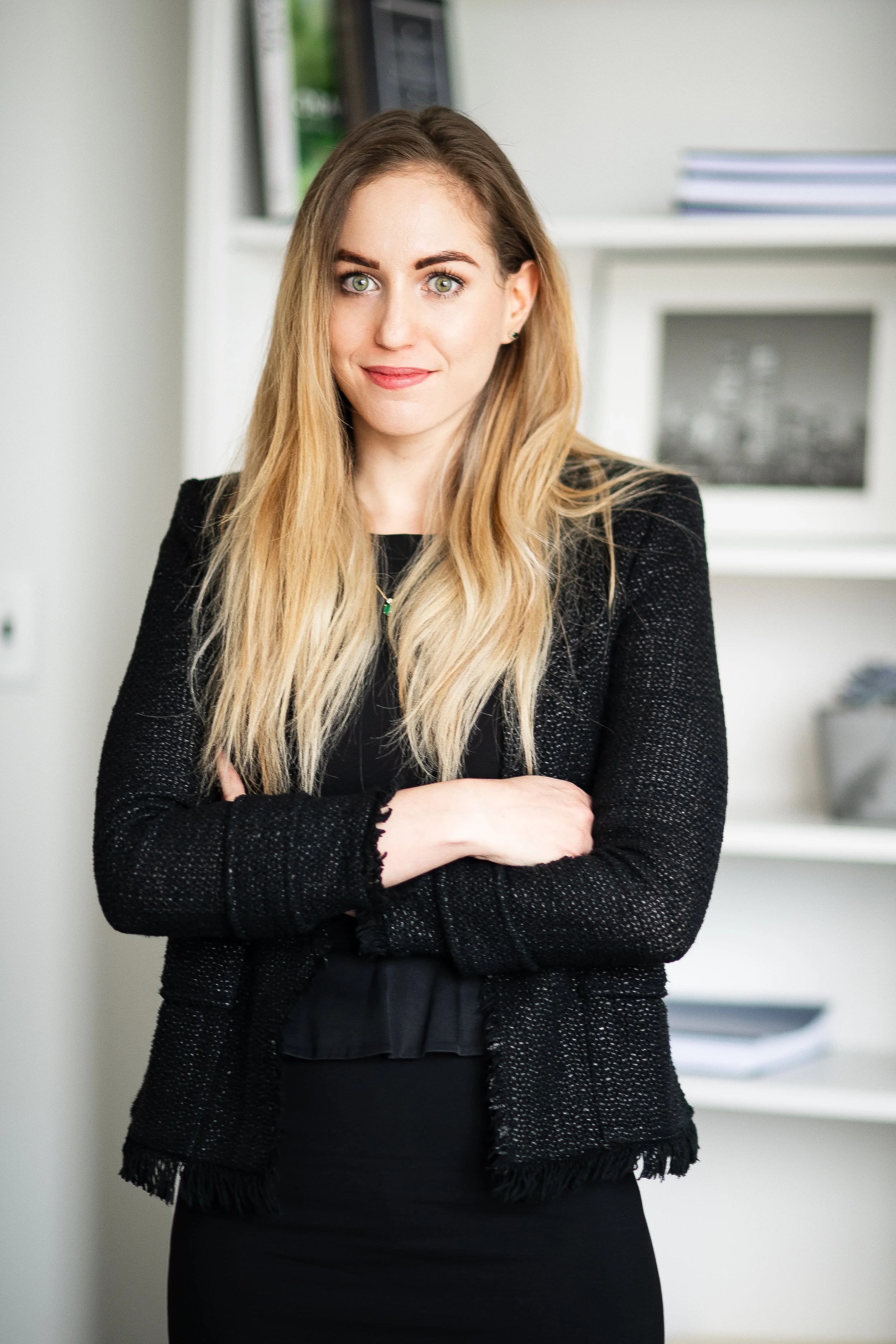 A woman with long blonde hair and green eyes standing with crossed arms in an office setting, wearing a black dress and black blazer.