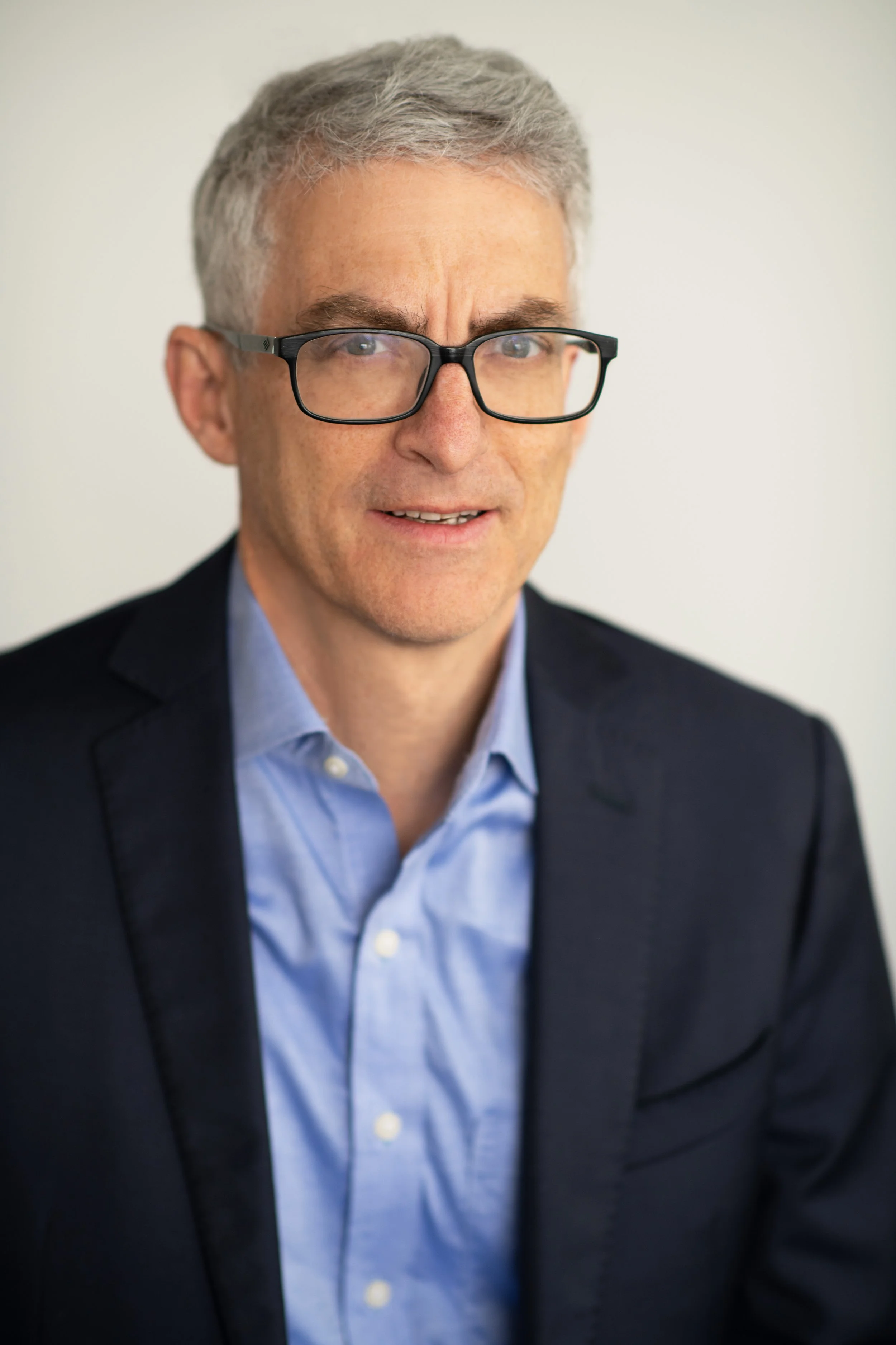 Close-up photo of a middle-aged man with gray hair, wearing black-rimmed glasses, a light blue shirt, and a dark blazer, against a plain background.