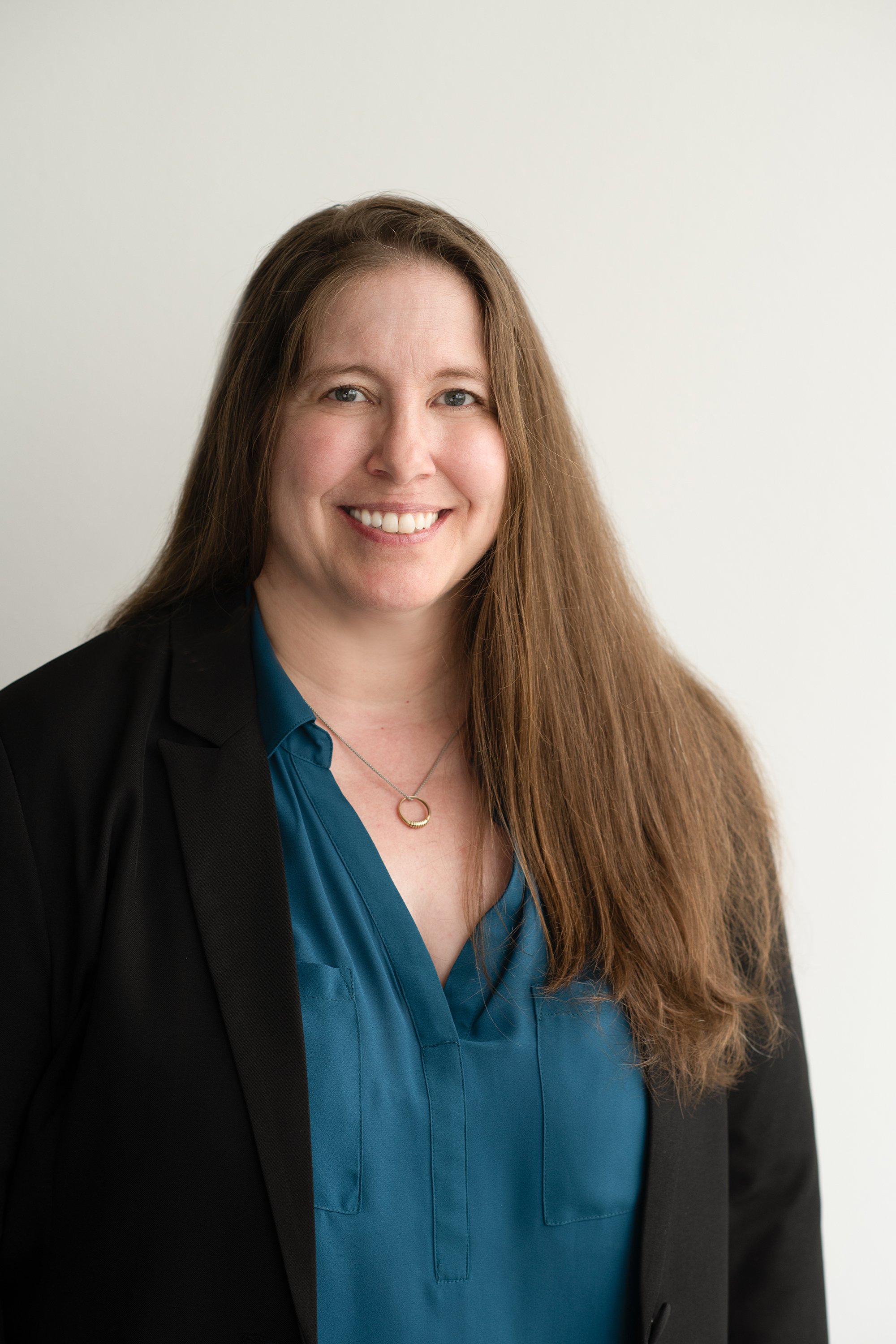 A woman with long brown hair, smiling, wearing a blue blouse and a black blazer, standing against a plain white wall.