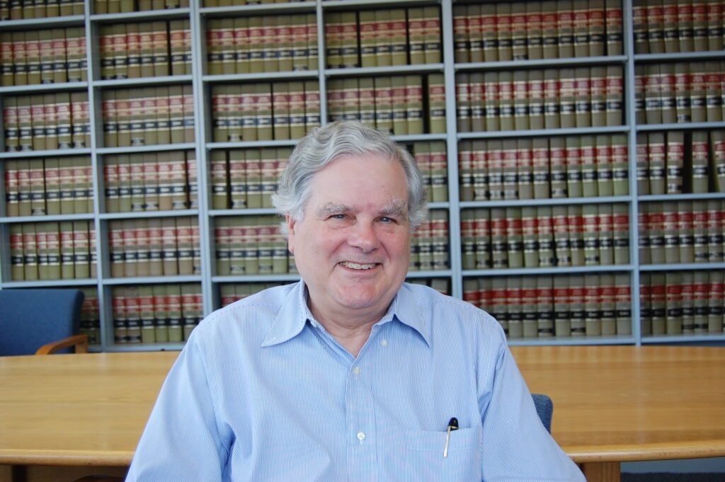 A smiling older man with gray hair, wearing a light blue button-up shirt, sitting at a large wooden table in front of a wall of law books in a library or office.