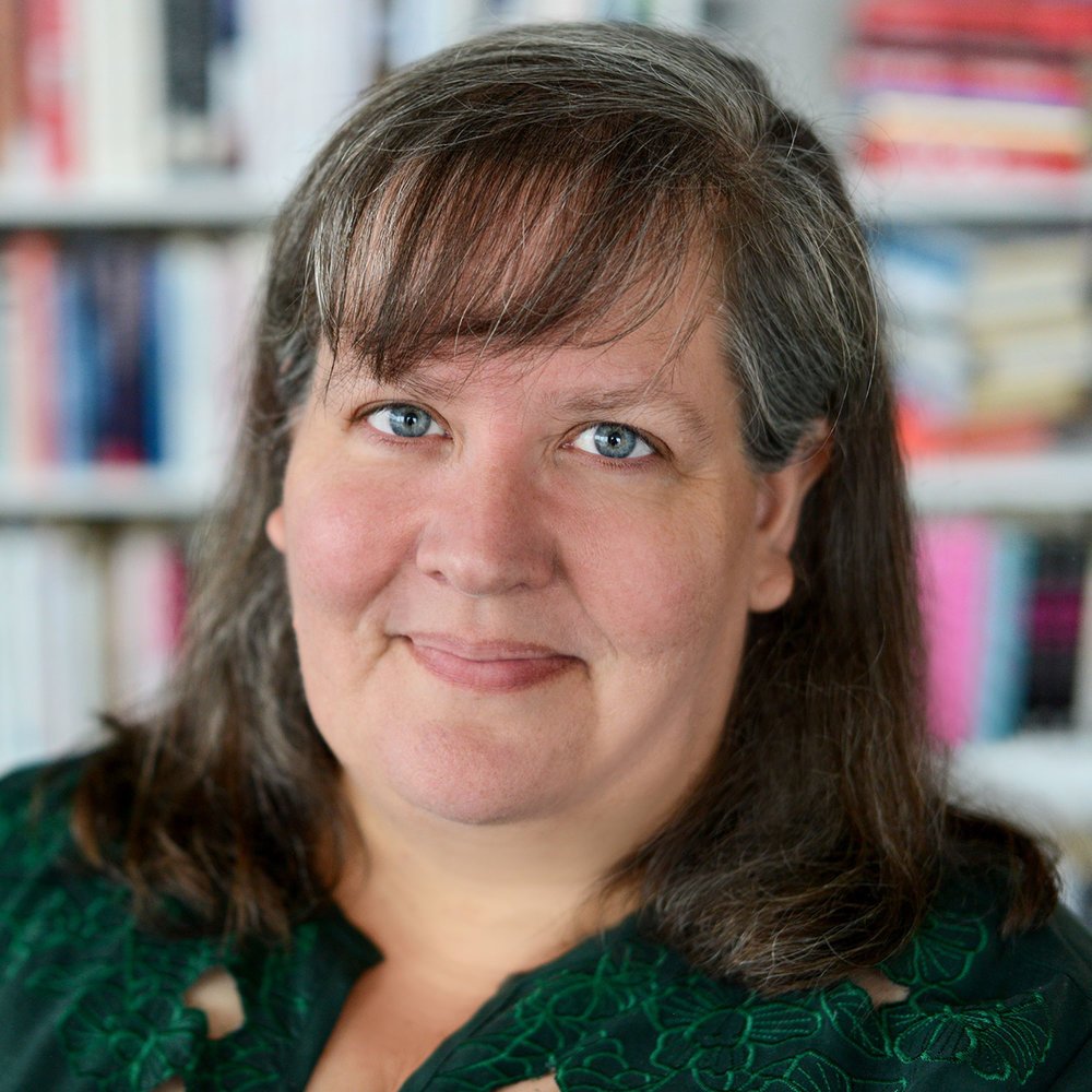 A woman with shoulder-length brown hair and blue eyes smiling, with bookshelves blurred in the background.