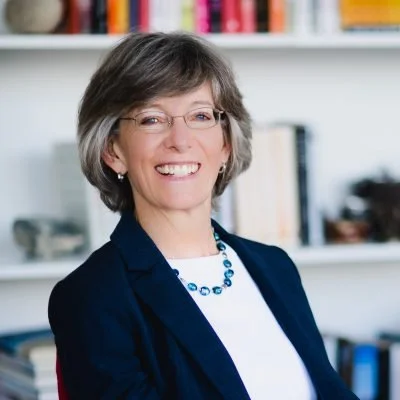A smiling woman with short gray hair and glasses, wearing a navy blazer and a blue necklace, standing in front of a bookshelf.
