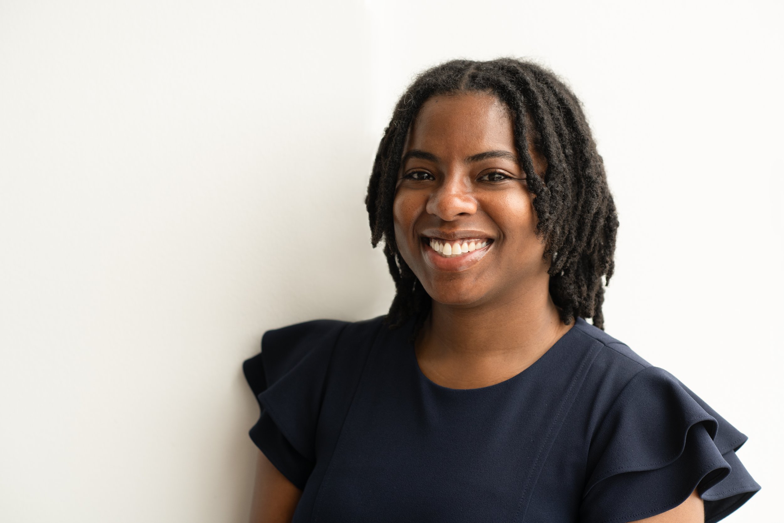 A smiling woman with dark hair styled in dreadlocks, wearing a dark-colored top with ruffle sleeves, standing against a white wall.
