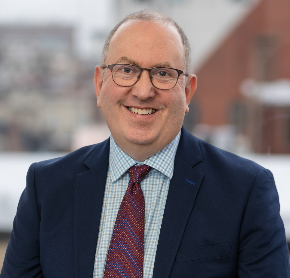 A smiling man with glasses, wearing a navy blazer, a checkered shirt, and a red tie, standing in front of a blurred background of buildings.