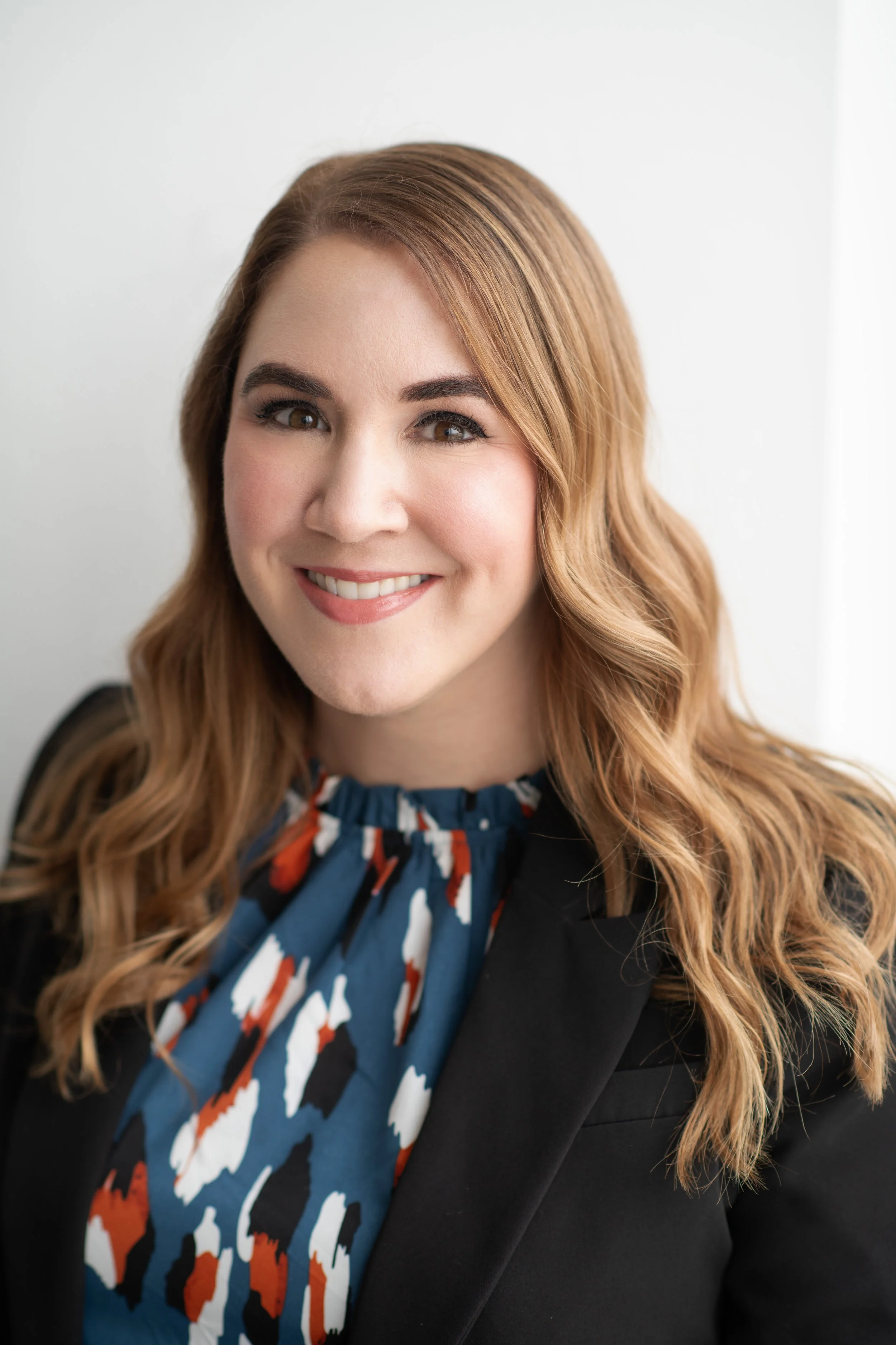 Close-up of a woman with long, wavy, light brown hair, smiling with visible teeth, wearing a patterned blue, red, orange, and white blouse and a black blazer.