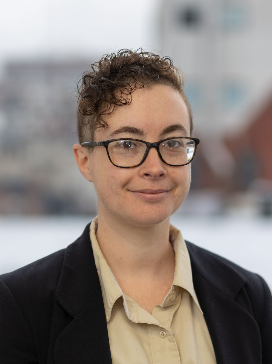 A woman with short curly hair, glasses, and a slight smile, wearing a beige shirt and black blazer, standing outdoors with blurred buildings in the background.